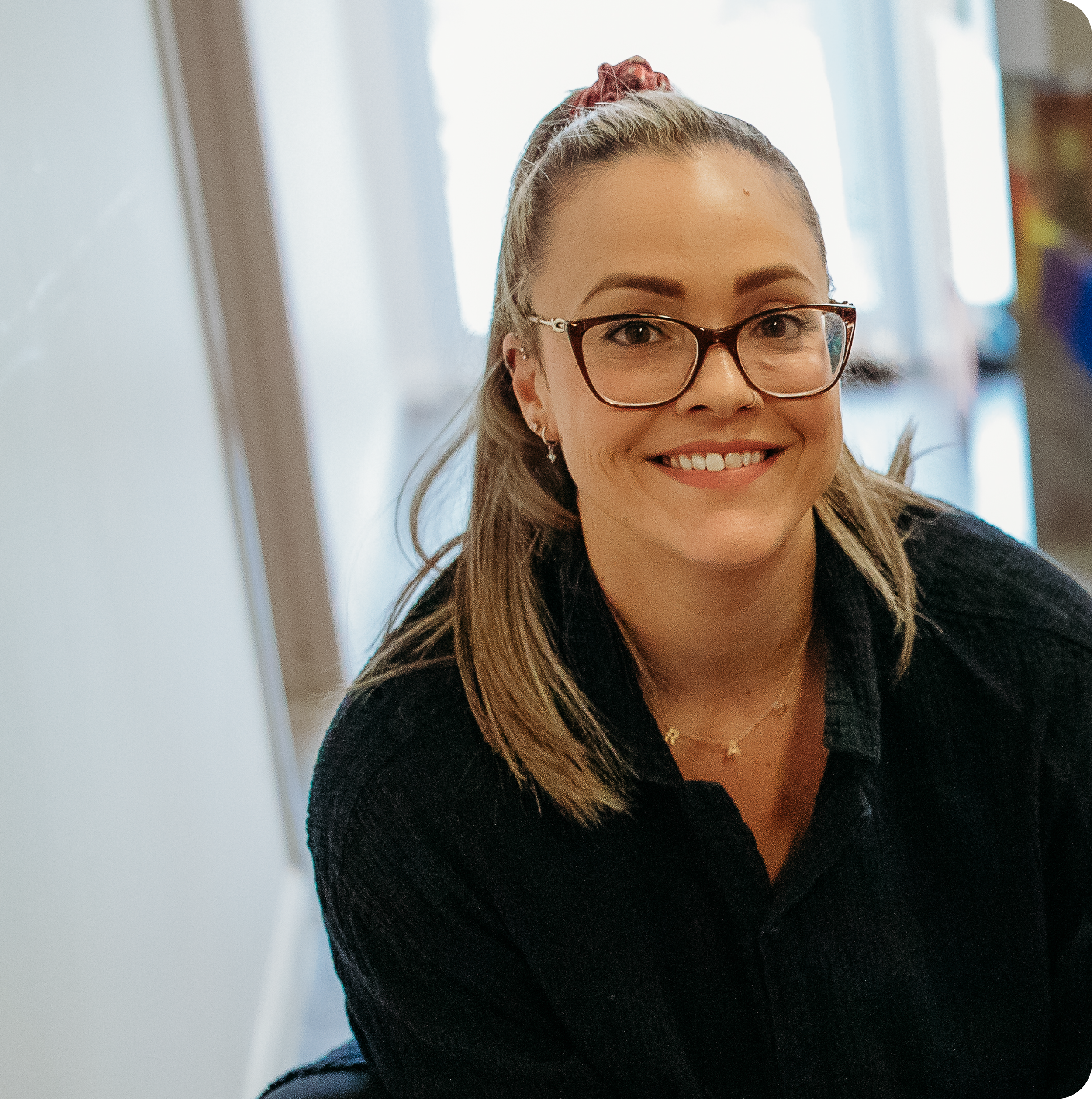 Close-up of a smiling woman with glasses and long, light brown hair, with a bun secured by a red scrunchie, in a bright indoor setting.