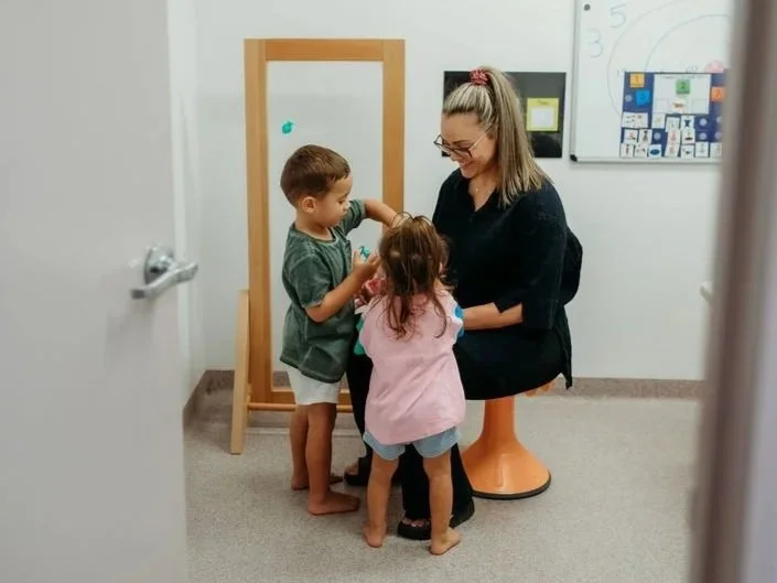 A woman sitting on an orange stool with two children, a boy and a girl, in a classroom or therapy room setting. The woman appears to be engaging with the children, who are standing close to her and each other. There is a whiteboard with colorful charts and a wooden-framed mirror or easel in the background.