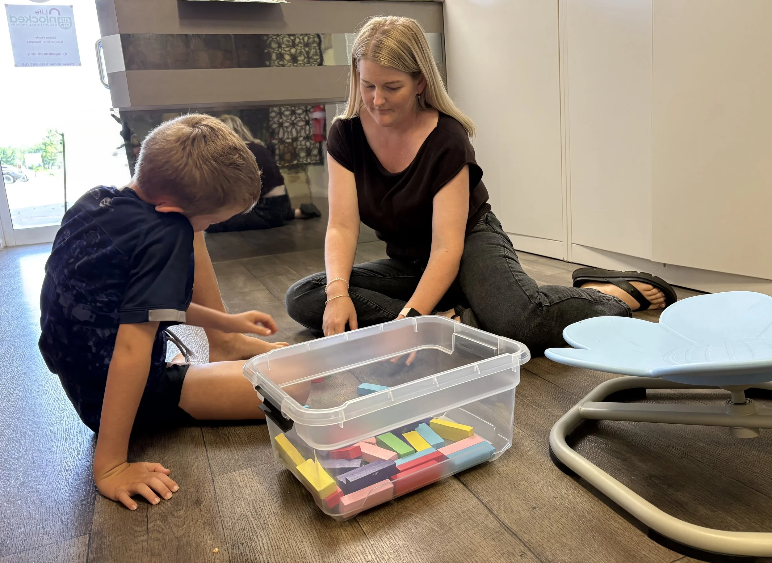 A woman and a young boy are sitting on the floor playing with colorful foam blocks; the woman is watching the boy, who is focused on stacking the blocks. There are additional foam blocks on a small table and in a plastic container nearby. The setting appears to be a children's play area or a classroom.