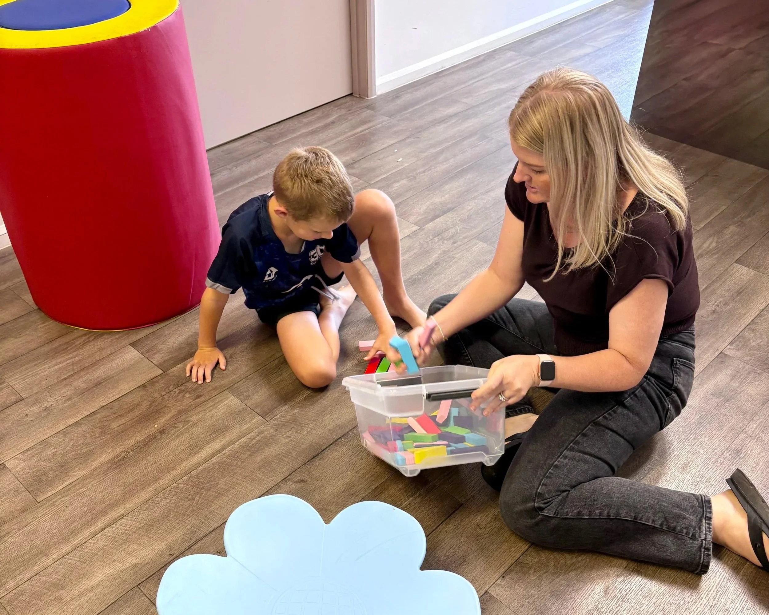 A woman sitting on the floor with a boy, playing with colorful domino tiles stored in a clear plastic container. Two round blue tables are in the foreground, and a red cylindrical object is partially visible on the left.