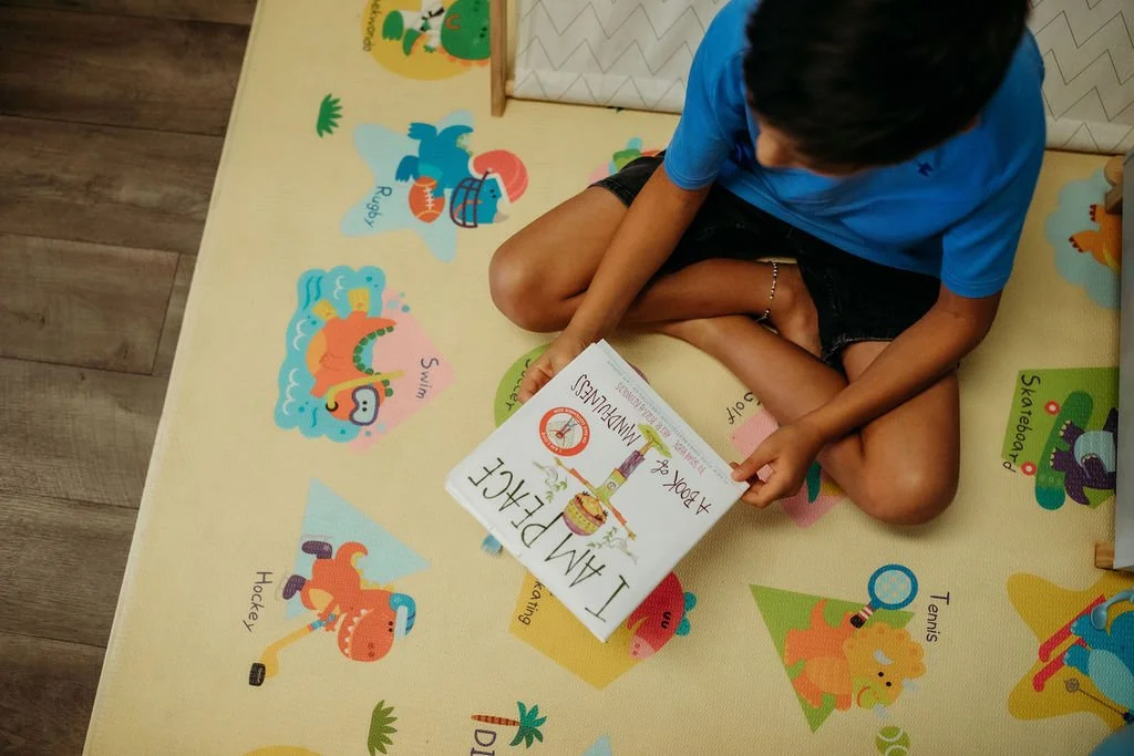 A boy sitting cross-legged on a colorful floor mat with illustrations of various sports and activities, reading a children's book titled 'I am Dairy-Free & Egg-Free'.