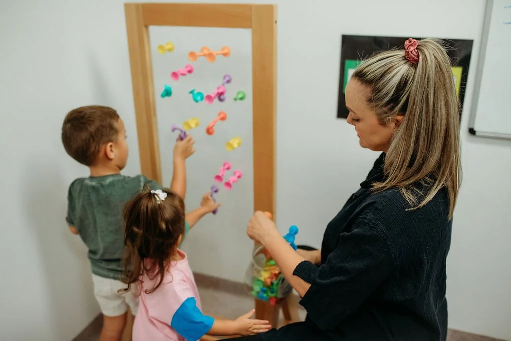 Two children and a woman playing with colorful magnetic fishing toys on a whiteboard in a classroom.