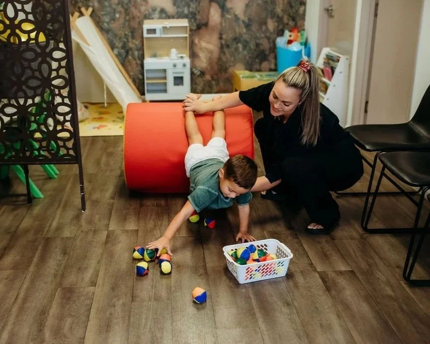 A woman helping a young boy who is crawling on the floor to pick up colorful toys from a basket in a playroom.