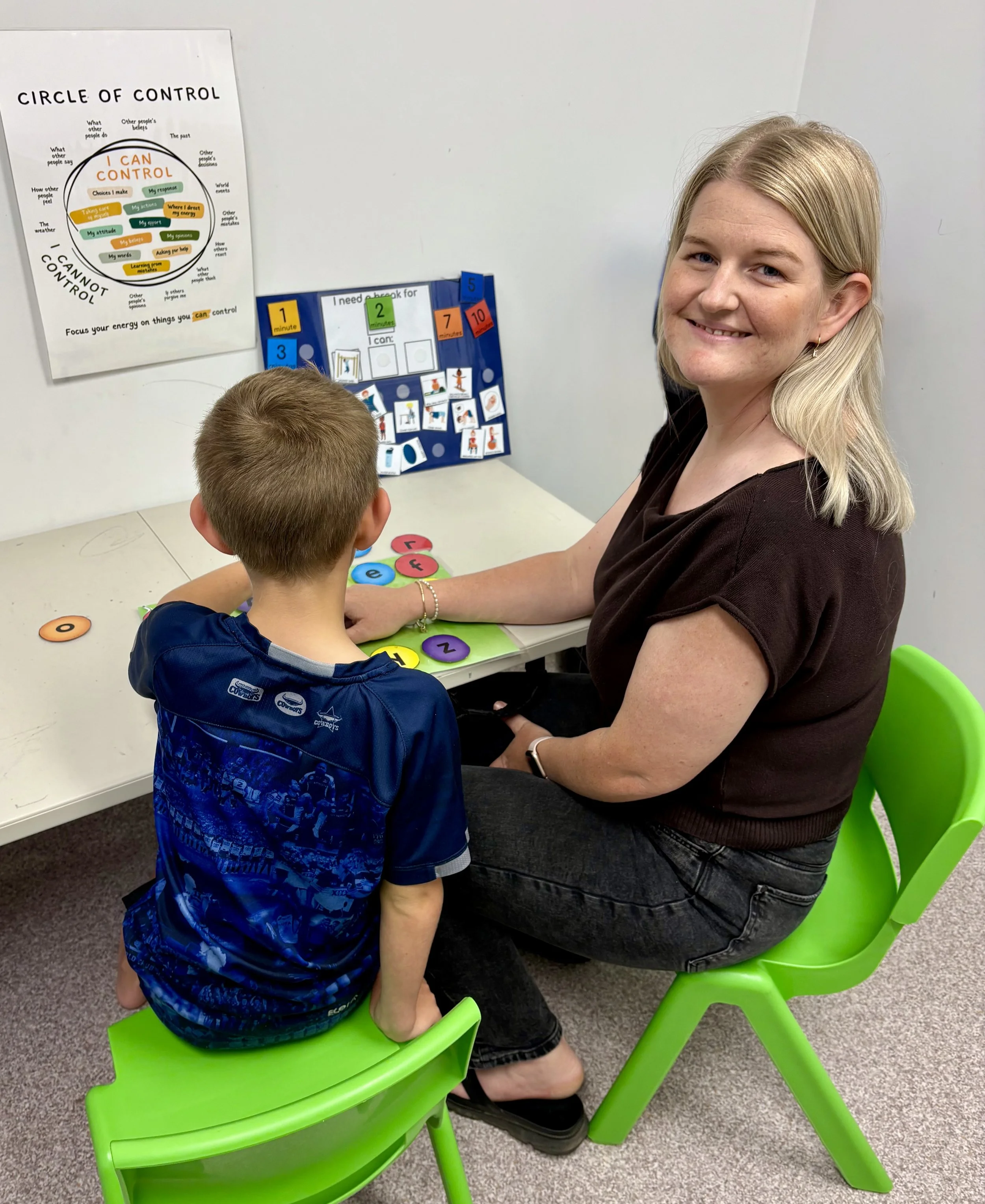 A woman sitting on a bright green chair at a desk with a young boy across from her. The woman is smiling and has blonde hair, while the boy has light brown hair and is facing away from the camera. On the wall behind them are educational posters, including one titled 'Circle of Control' and another with color-coded task labels.