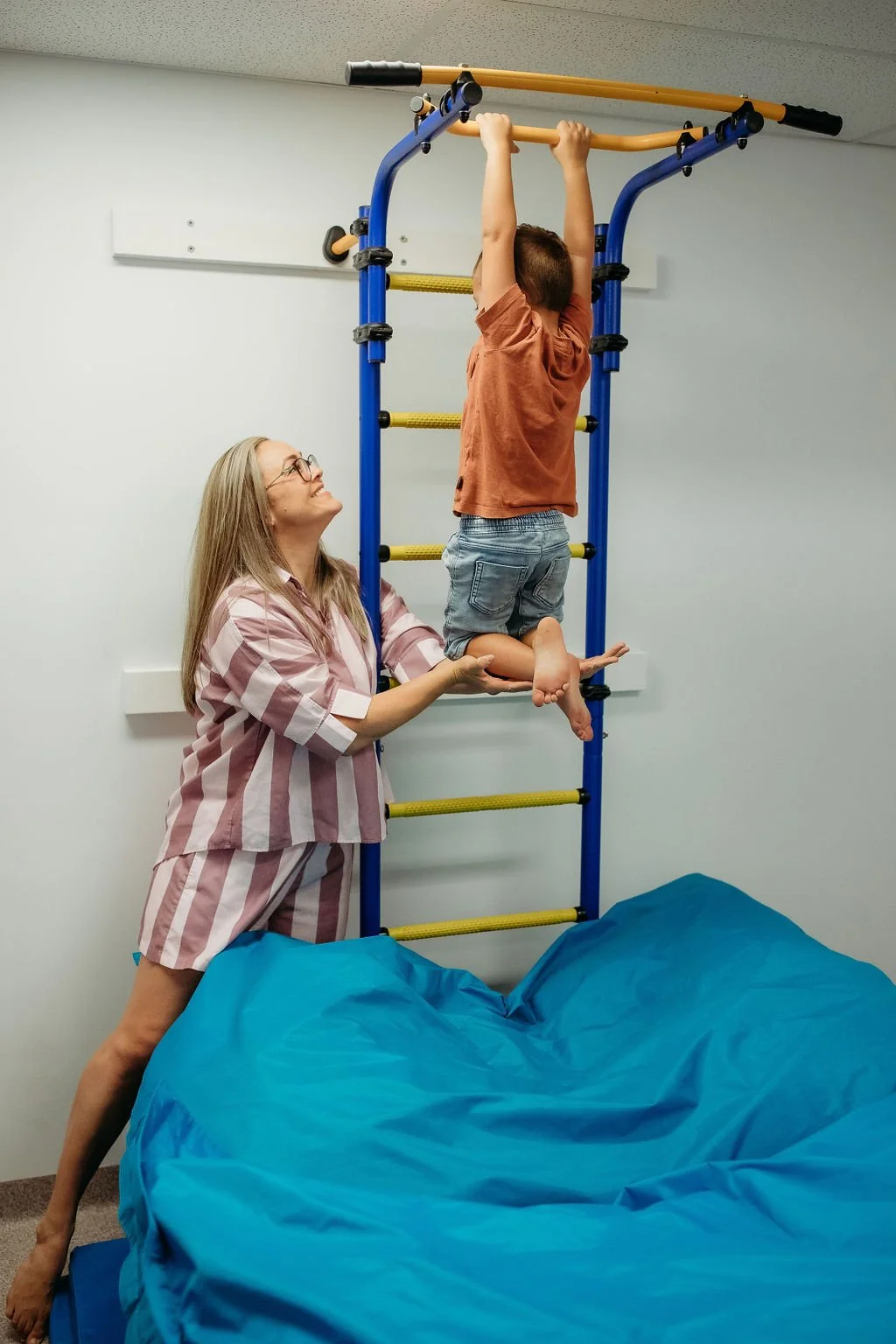 A child wearing a brown shirt and denim shorts is hanging from a yellow pull-up bar, supported by a woman in pink and white striped pajamas who is holding his legs. The child is upside down with arms extended upward grasping the bar. The setting appears to be an indoor play or therapy room with a padded blue mat on the floor.