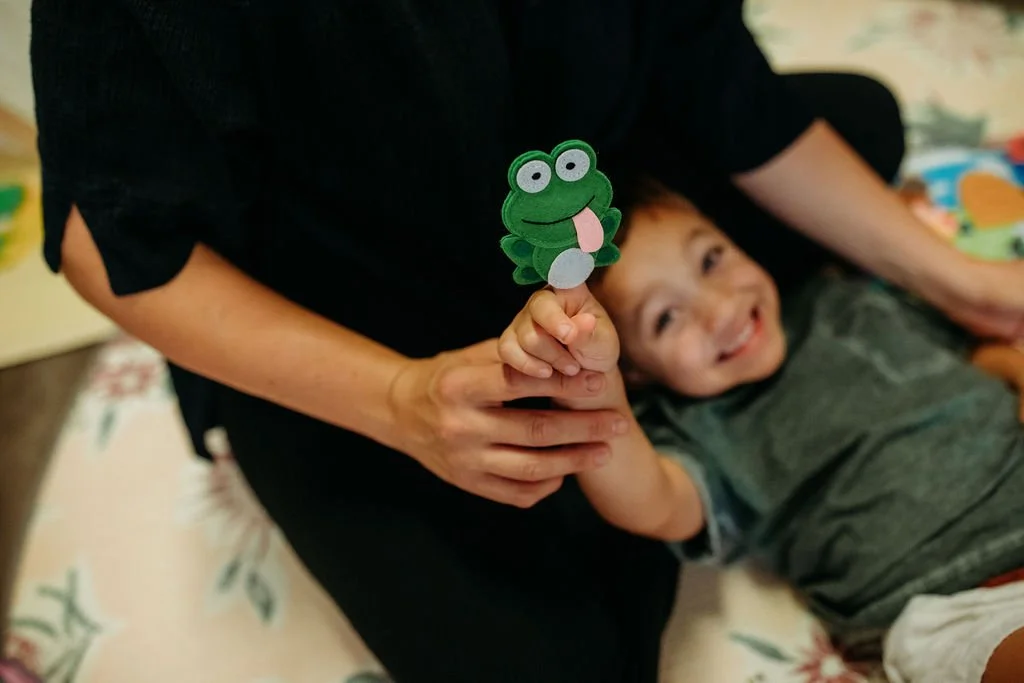 A child lying down and smiling, holding a frog-themed puppet, with an adult holding the puppet's hand.