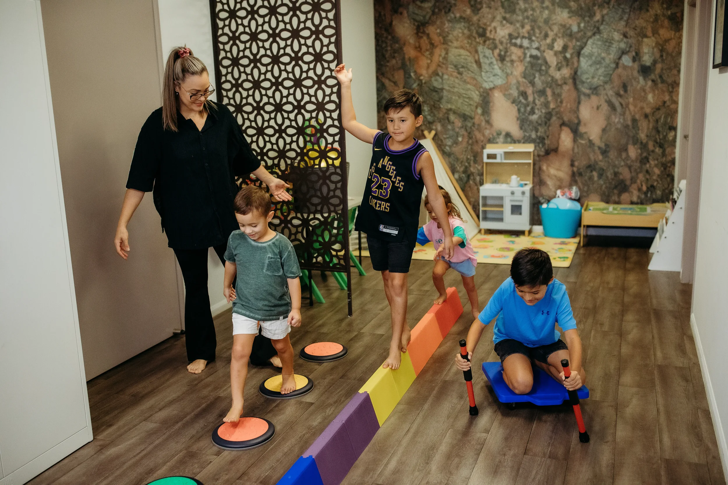 A woman guiding children in an indoor activity, balancing on colored stepping stones, with some children seated in a small blue wagon, against a background of a decorative wall and play area.