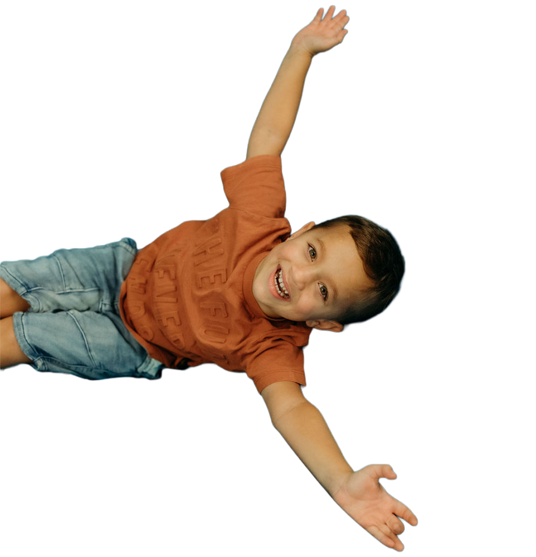 Smiling young boy in orange t-shirt and denim shorts lying on the floor with arms extended outward.