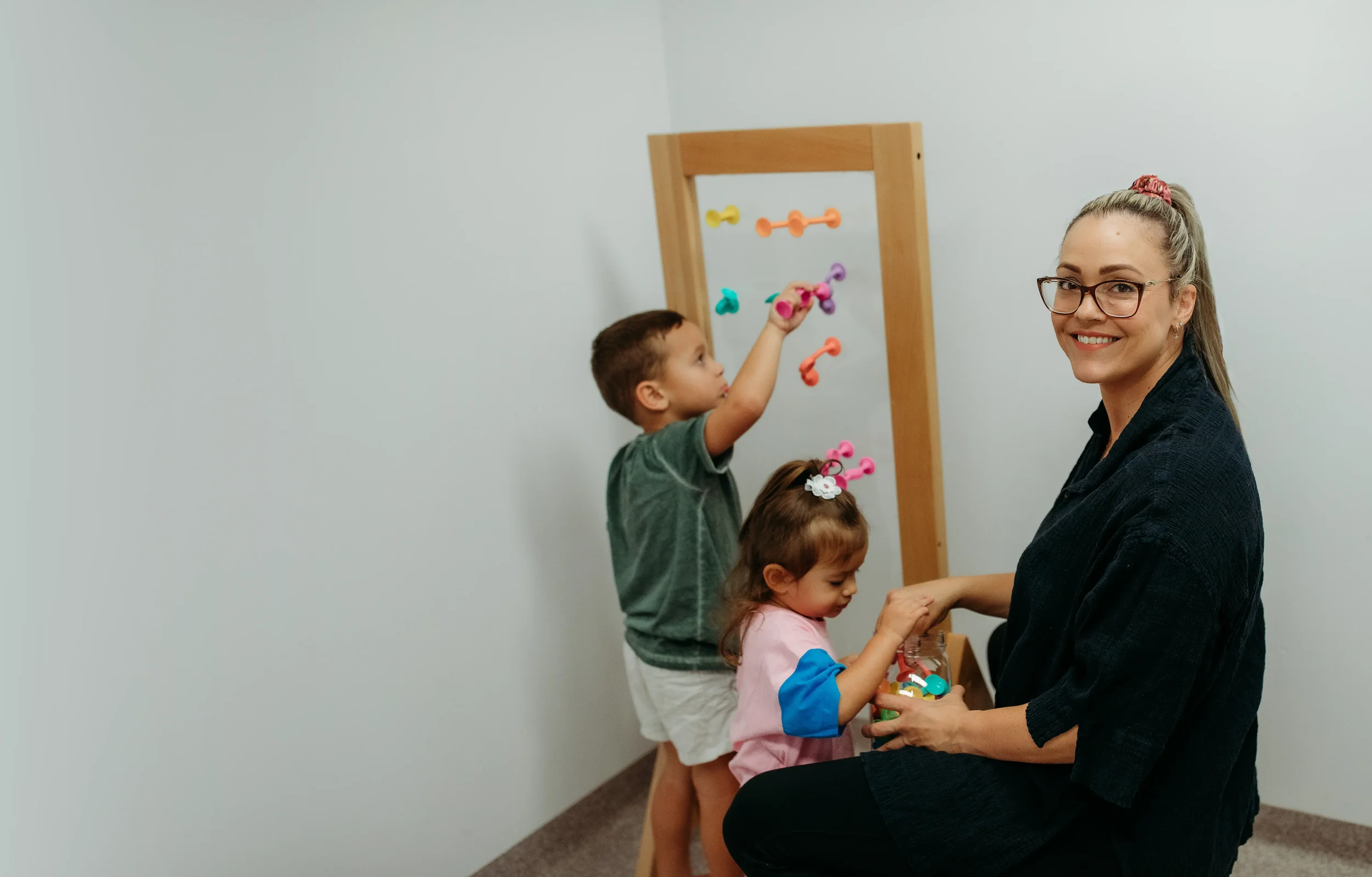 A woman with glasses and a ponytail smiling while holding hands with a young girl and surrounded by children in front of a wooden activity frame with colourful suction toys.