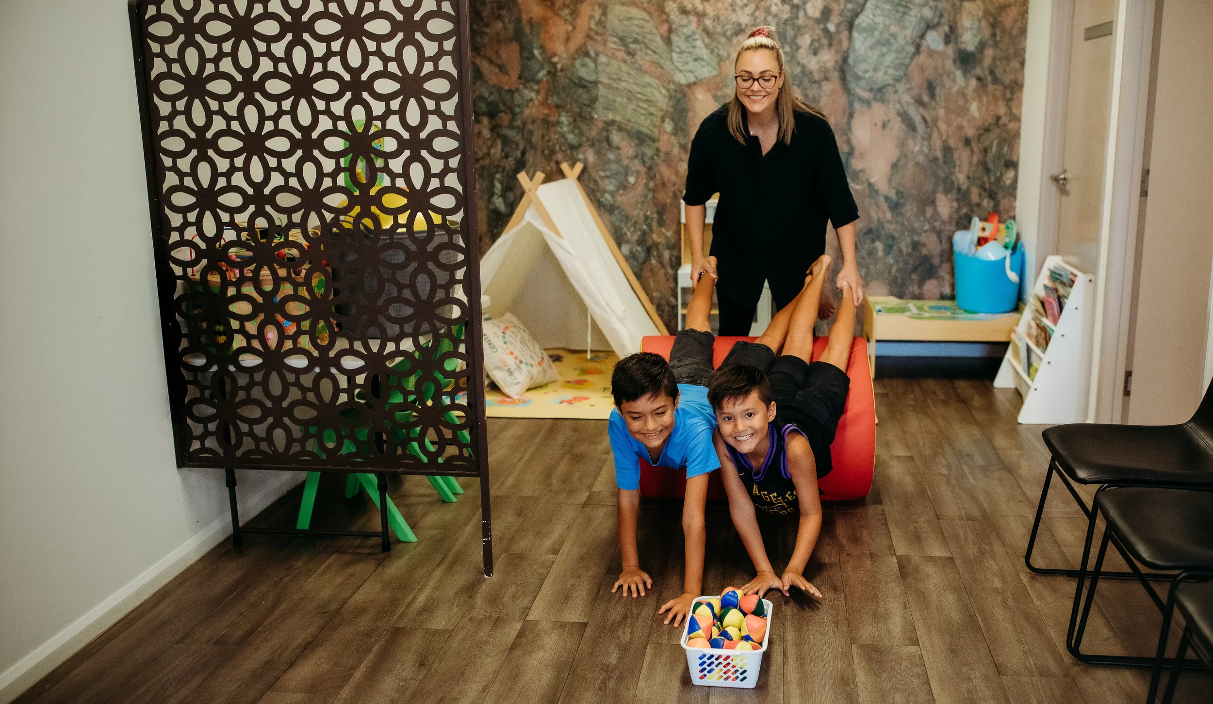 A woman and two children playing a game together inside a room, with a small tent and a decorative partition in the background.