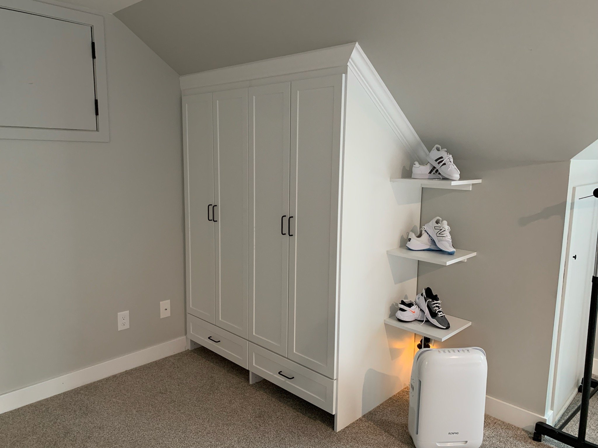 White storage cabinet next to white wall with three black sneakers on white shelves and a white portable air purifier on carpeted floor.