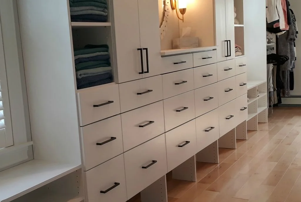 A modern, white storage cabinet with multiple drawers and open shelves, some with folded clothes, in a bedroom or walk-in closet with light wood flooring.