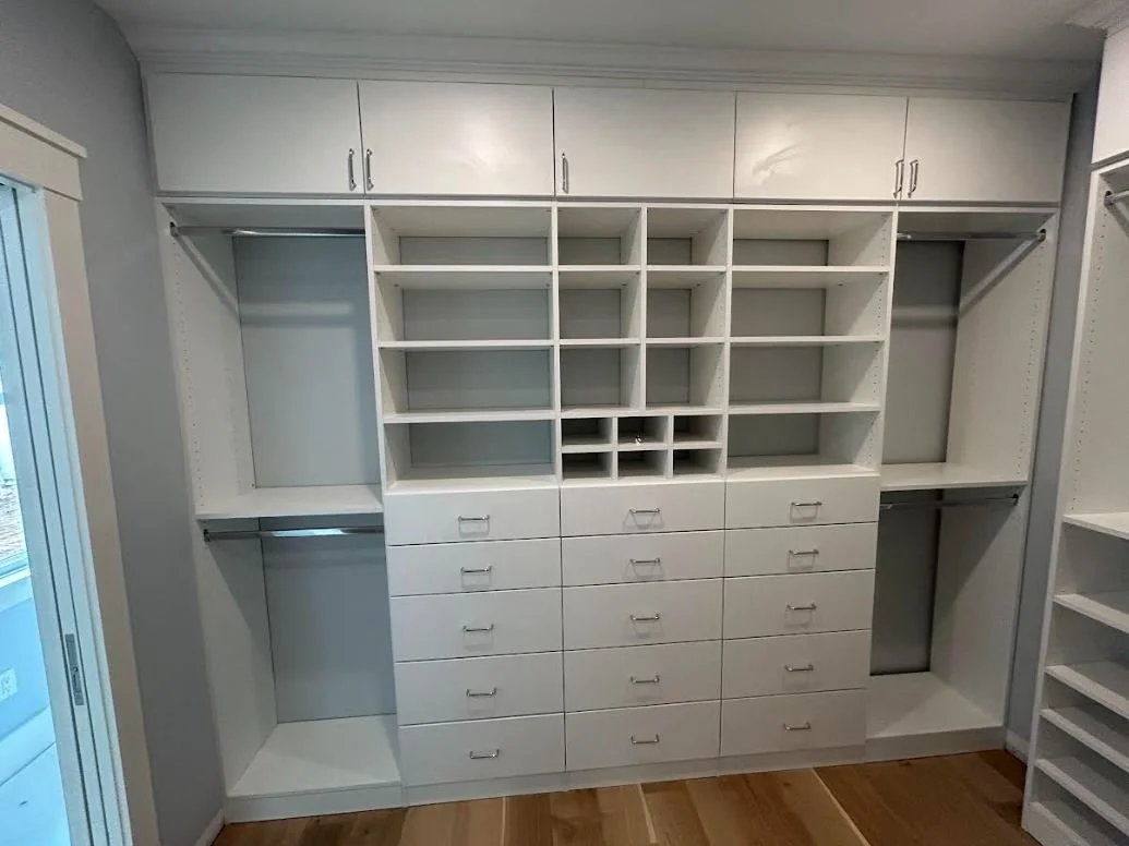 Empty white closet with multiple shelves, drawers, and hanging rods in a room with hardwood floor.