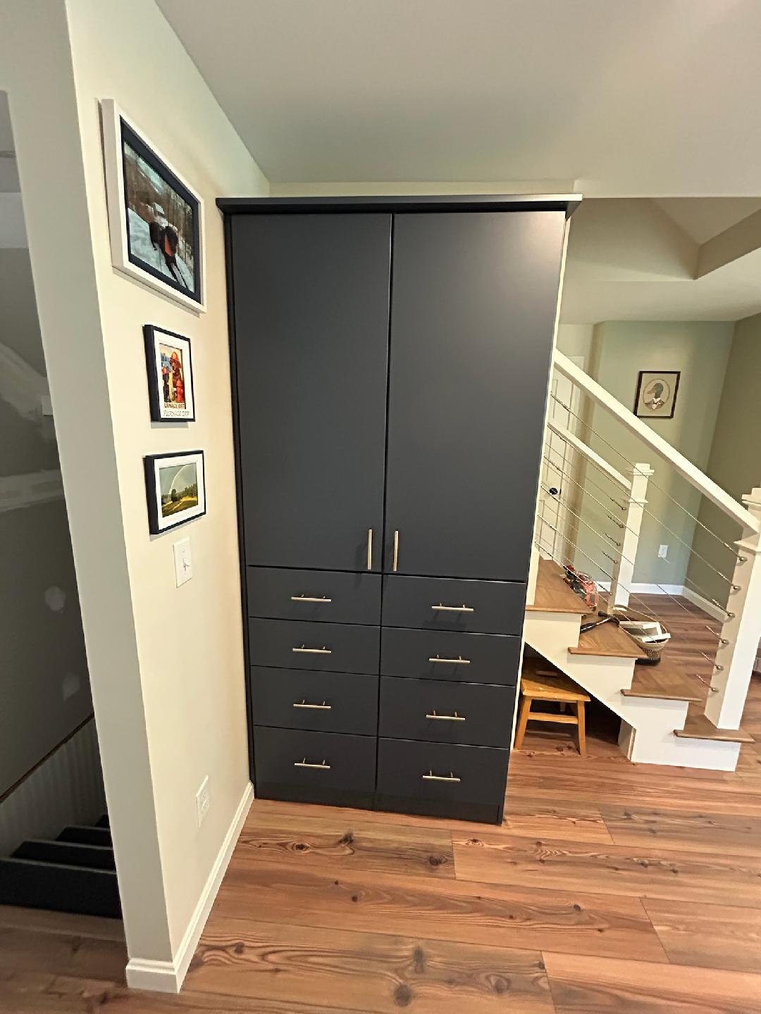 Dark gray cabinet with multiple drawers and double doors situated in a home interior near stairs with wooden flooring.