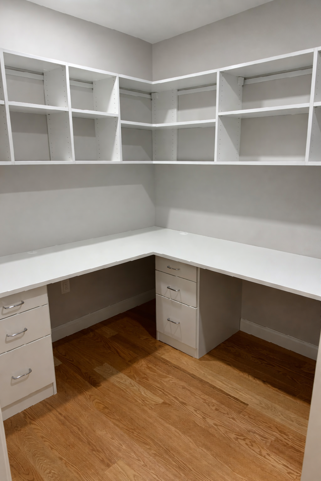 Empty white built-in corner desk with open shelving and drawers, hardwood floor.