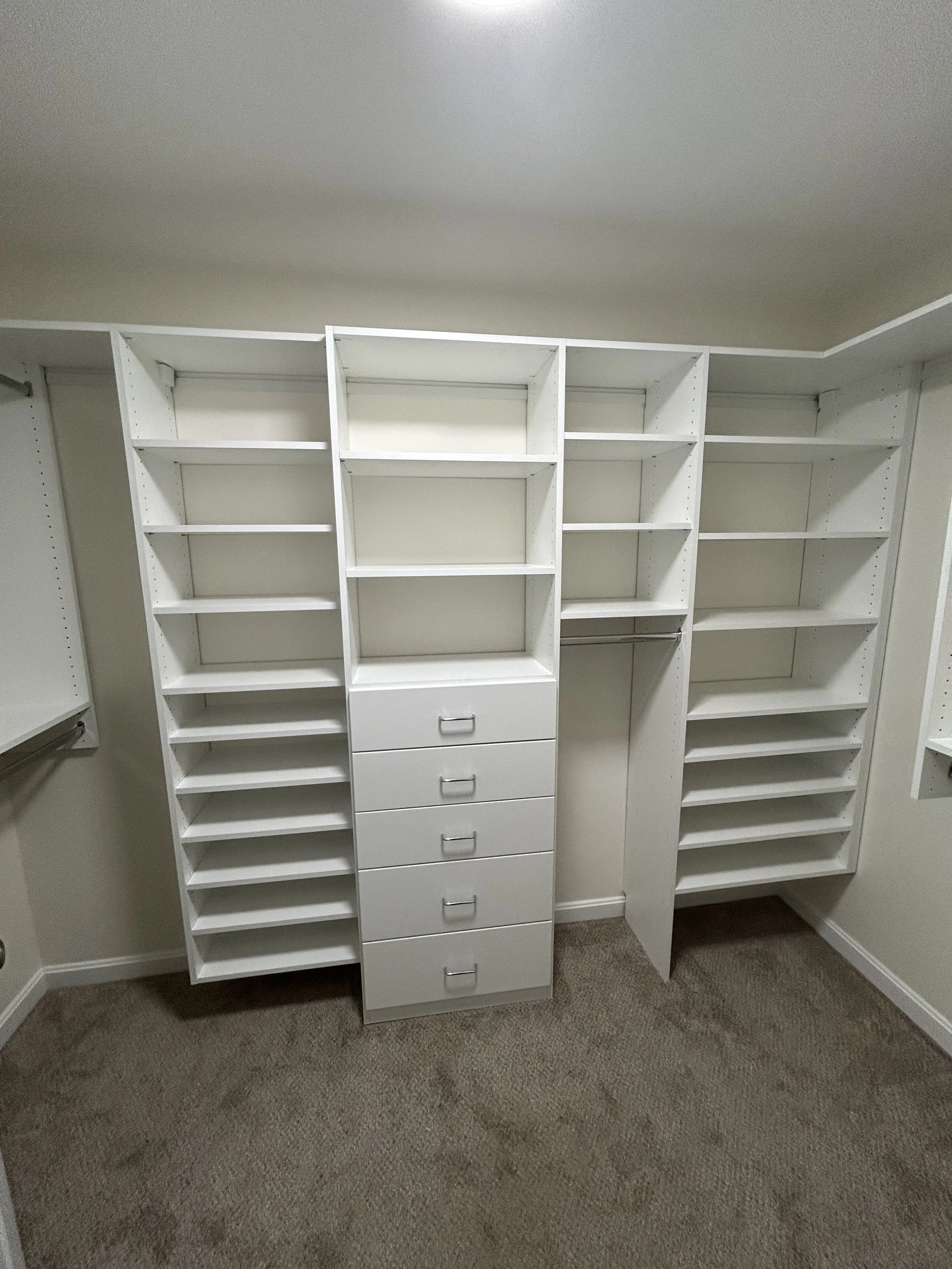 Empty white walk-in closet with shelves, drawers, and hanging rods, beige carpeted floor, overhead lighting.