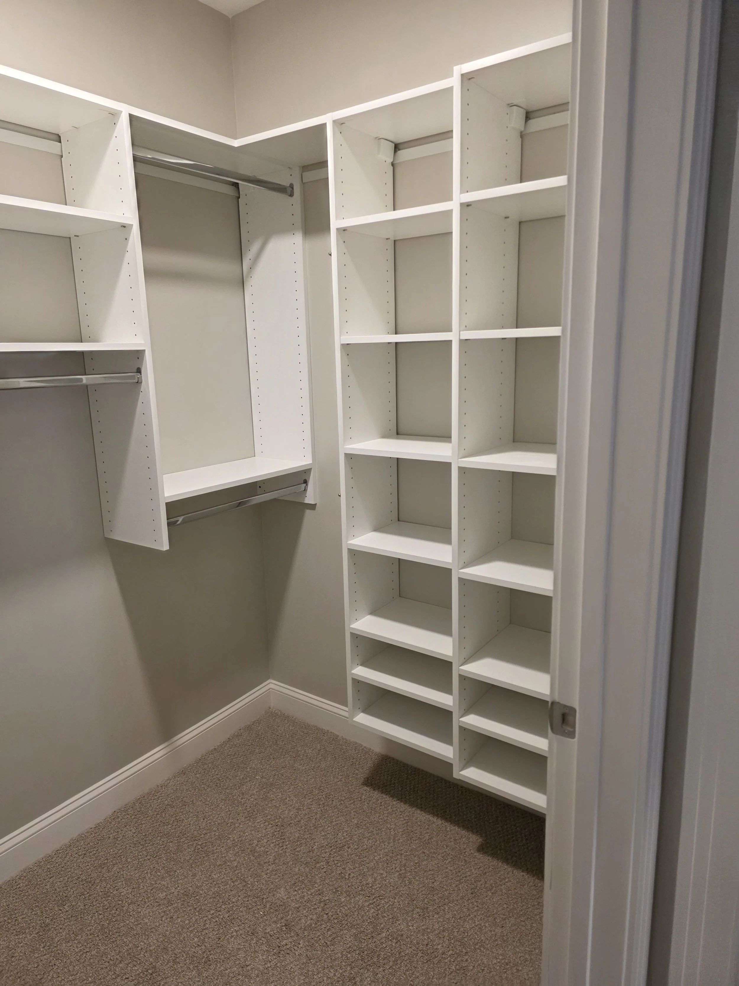 Empty small walk-in closet with white built-in shelves and hanging rods, beige carpet, cream-colored walls.
