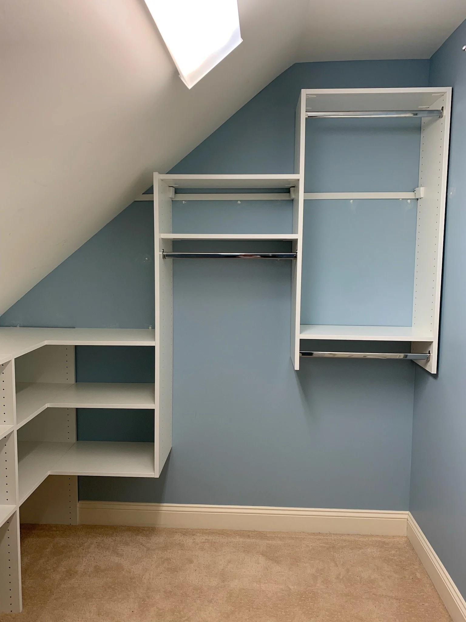 Empty custom closet with shelves and rods, white wood, against blue wall, in attic with sloped ceiling and carpeted floor.