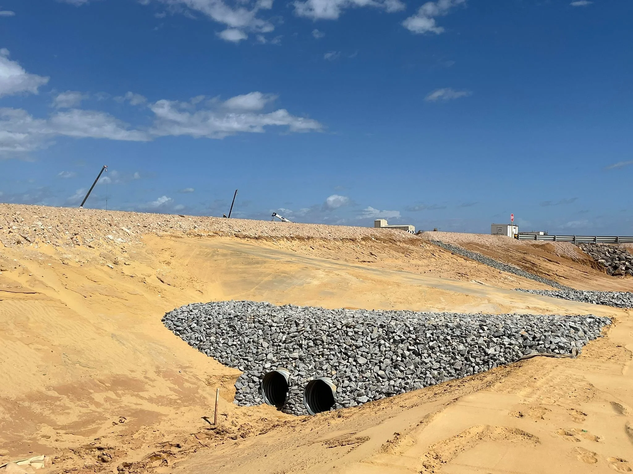 Construction site on sandy terrain with two large black culverts, gravel pile, and a clear sky with some clouds, with structures and equipment in the background.