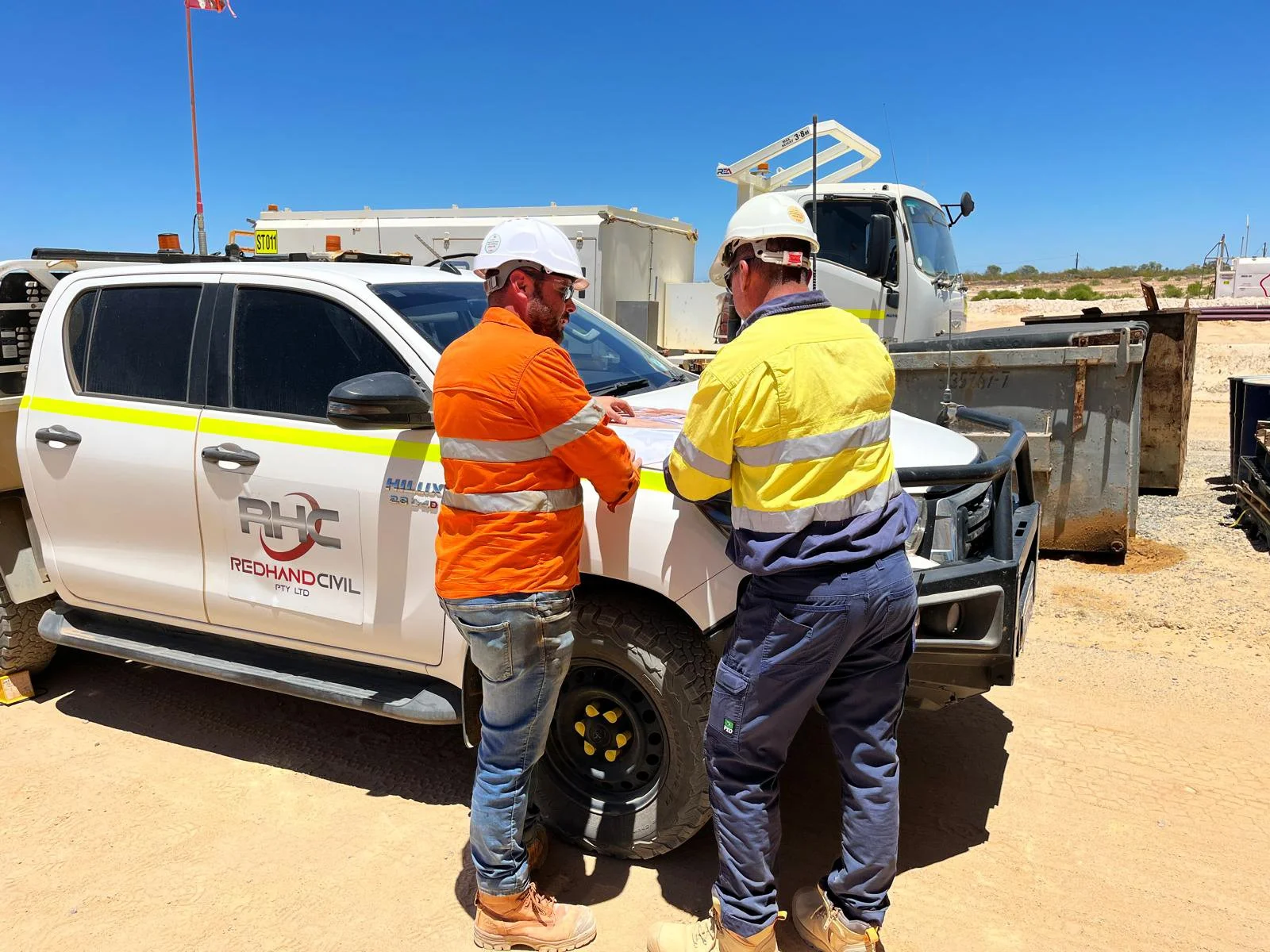 Two construction workers wearing safety helmets and reflective vests inspecting a white service vehicle on a construction site with clear blue skies.