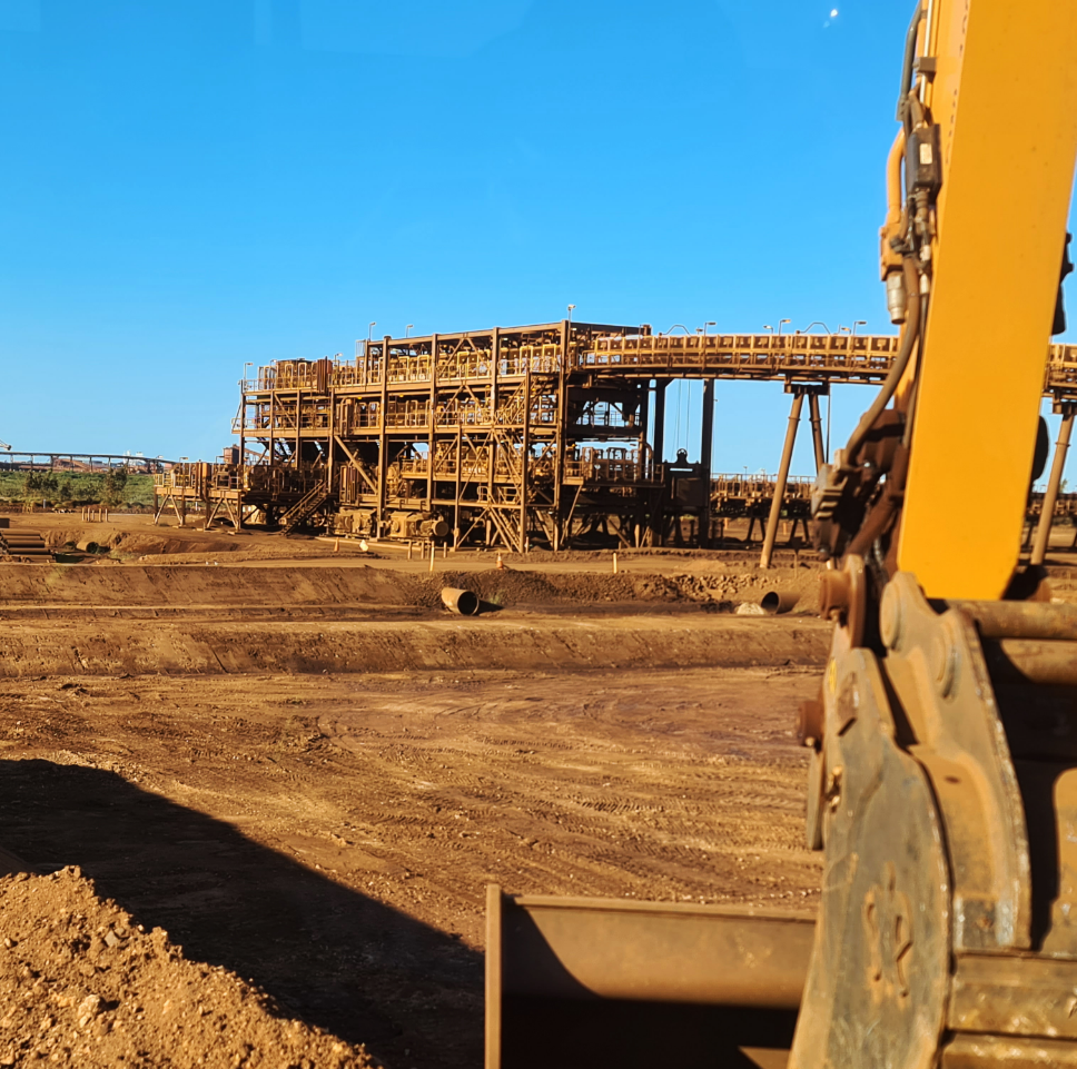 A construction site with a large industrial structure in the distance and a yellow excavator in the foreground under a clear blue sky.