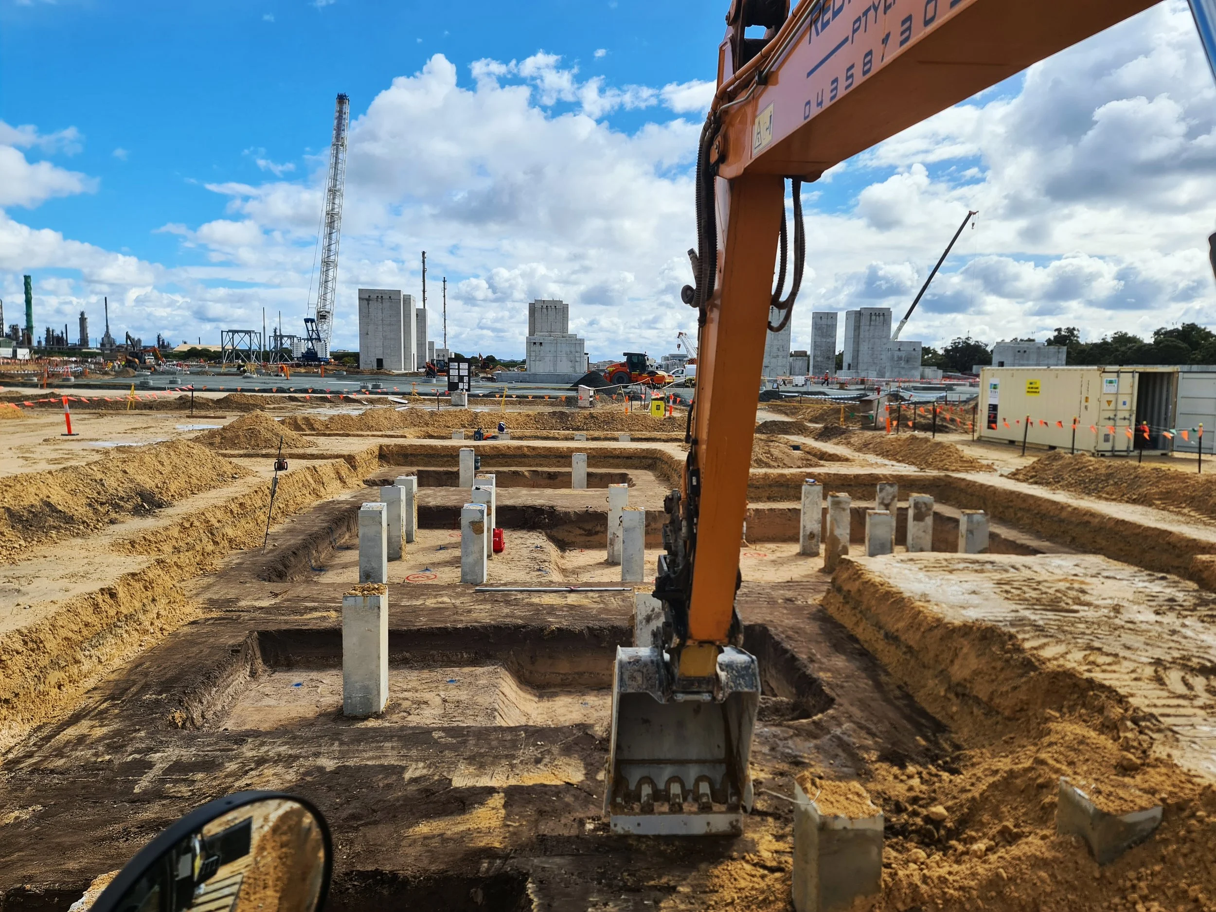 Construction site with ongoing excavation and foundation work, with a large crane and concrete structures in background, under a partly cloudy sky.