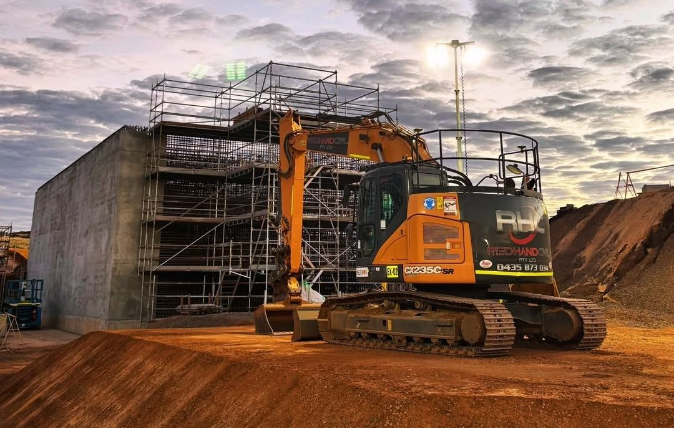 Construction site with a yellow excavator in front of scaffolding against a cloudy sky during sunset.
