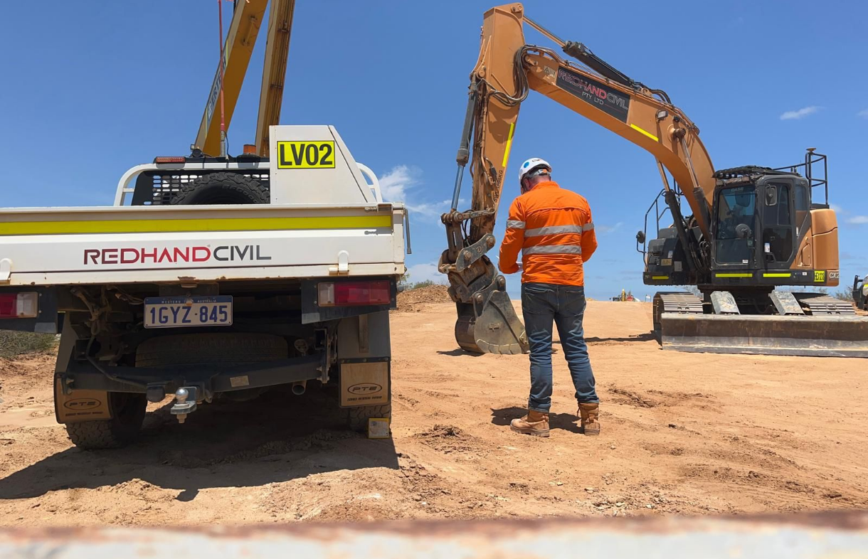Construction worker in an orange safety jacket and helmet standing next to a large excavator on a dirt construction site.