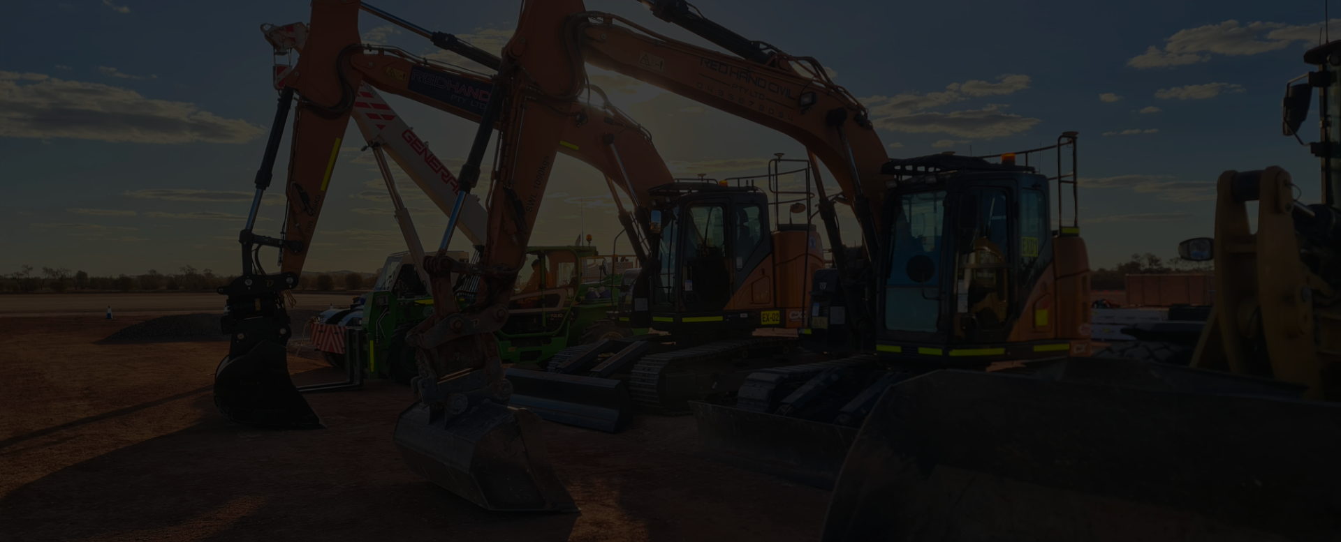 Construction site with large orange excavator operating on dirt under a partly cloudy sky at sunset.