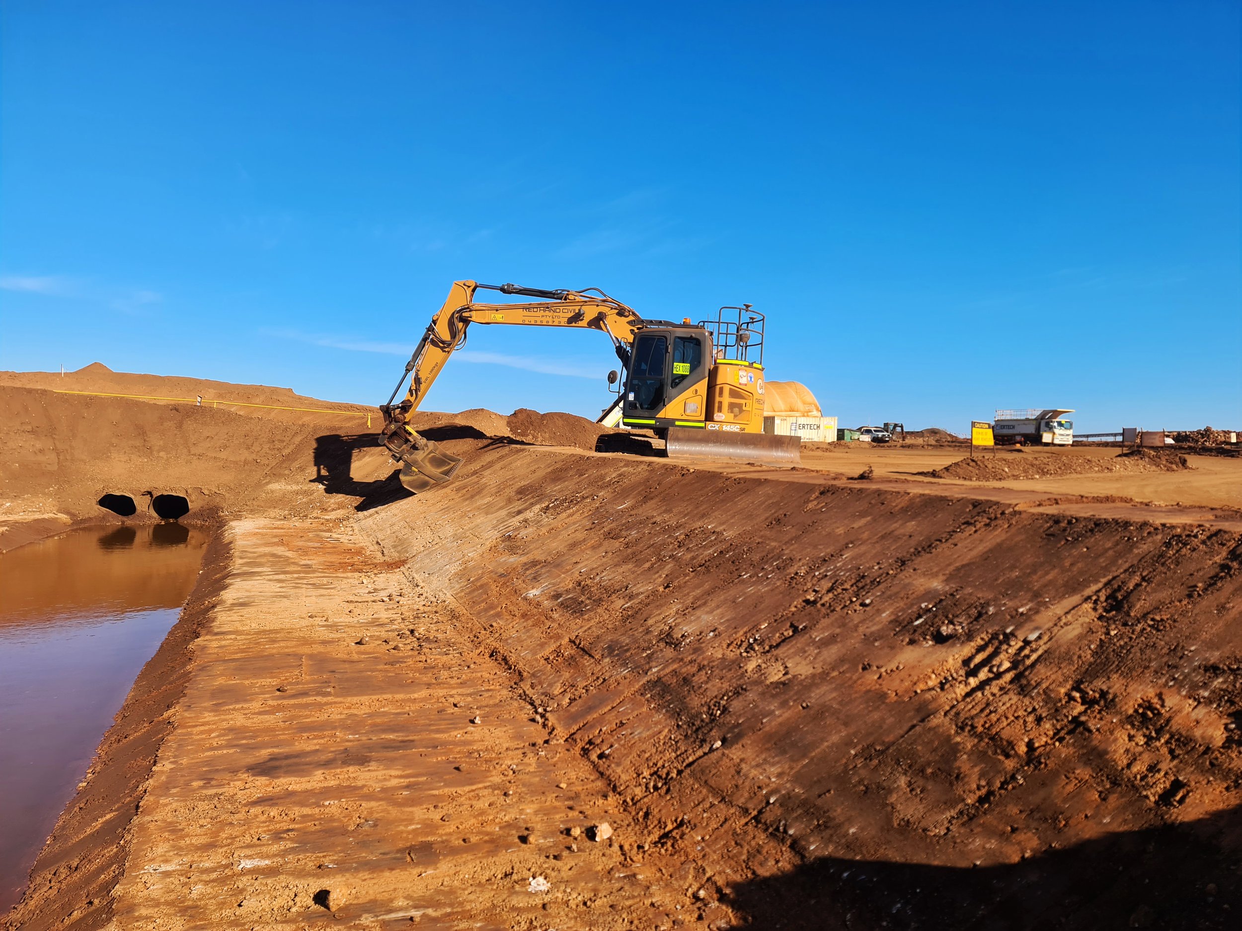 A yellow excavator working on a construction site near water under a clear blue sky.