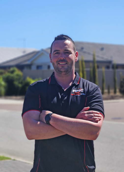 Emmet Harte - A man with short dark hair and a beard standing outdoors with arms crossed, wearing a black polo shirt with red accents and a logo, in front of a suburban neighbourhood in Western Australia.