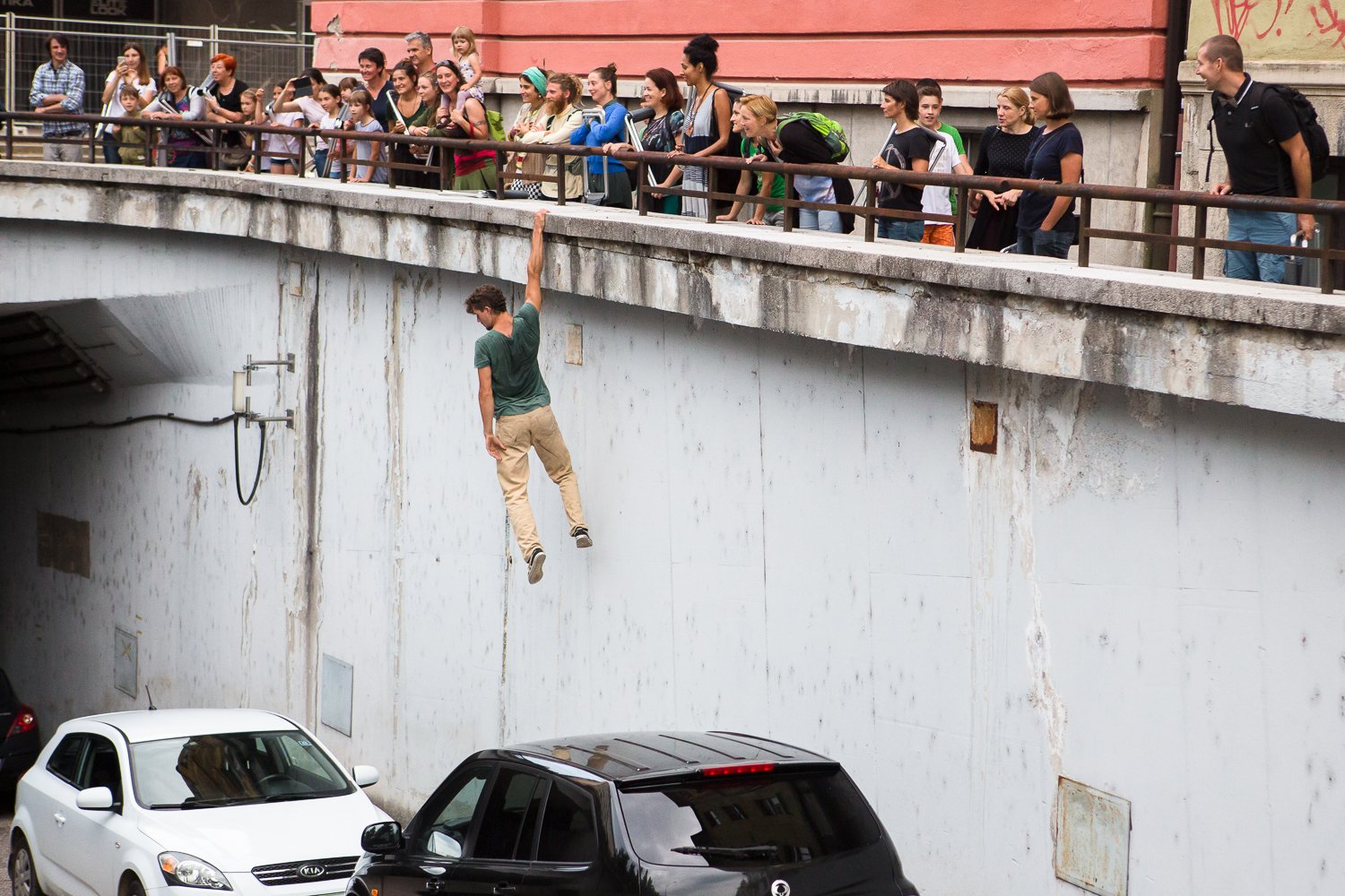 A man is hanging from the edge of a bridge over a parking lot, holding onto the rail, while a group of onlookers watches from above.