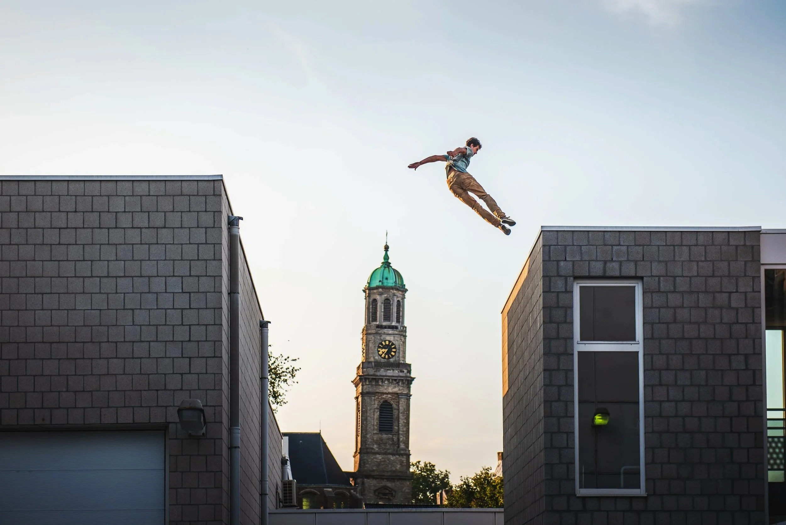 A person mid-air jumping between two buildings with a church steeple in the background.