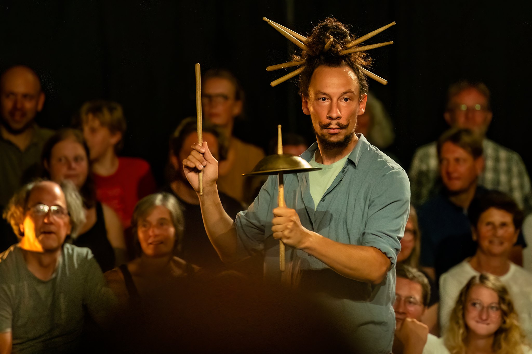 Man with lions mane styled hair in a blue shirt holding multiple drumsticks and a cymbal in a performance space, with an audience watching.