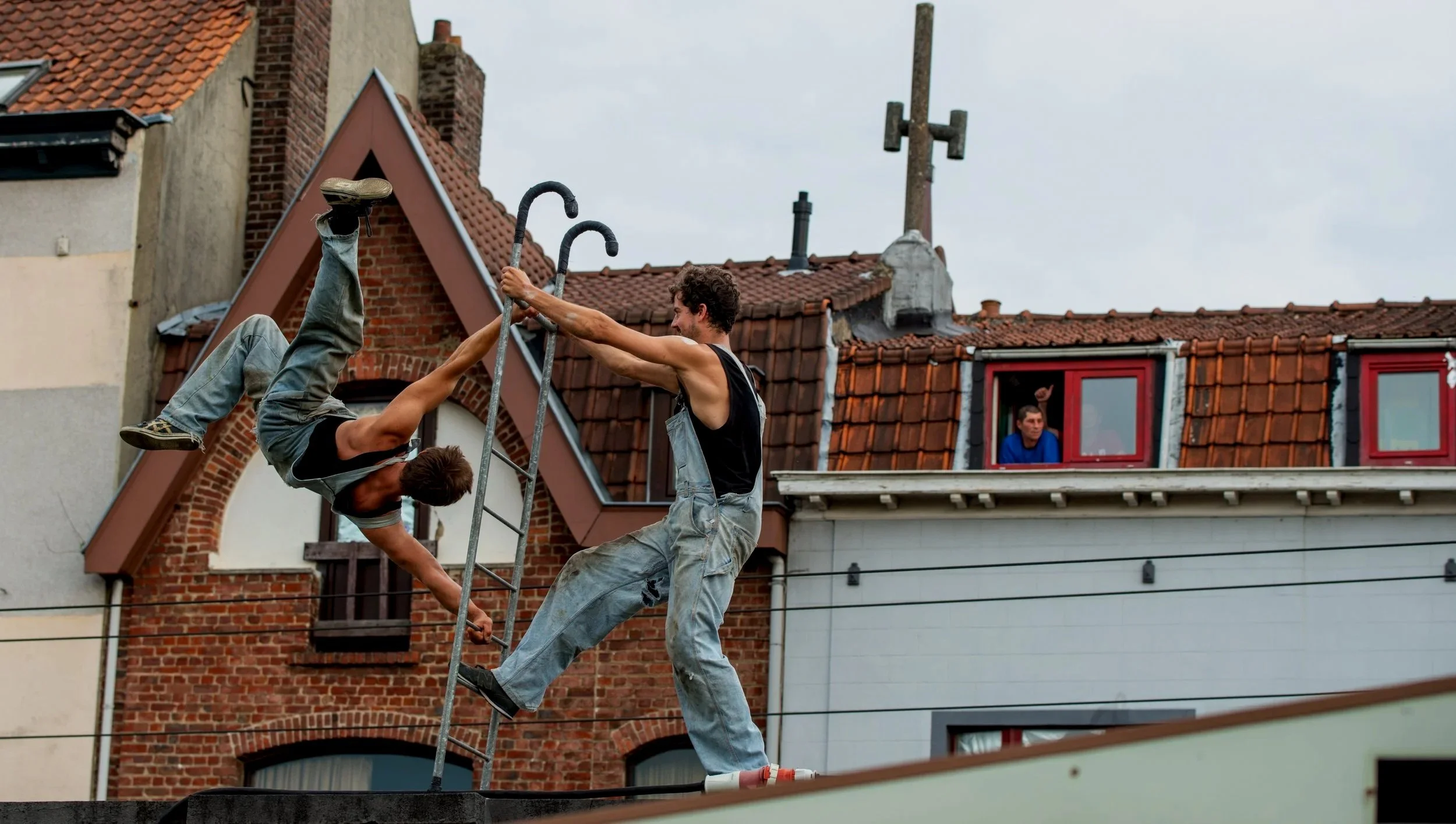 Two men practicing acrobatics on a rooftop with brick and stucco buildings in the background. One man is hanging upside down on a ladder, while the other is holding him by the arms. A person is watching from a window of the building in the background.
