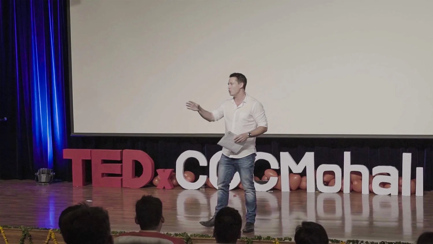 A man giving a presentation on a stage at a TEDx event in Cochin, Mangalore, with large pink and white letters spelling out 'TEDxCochinMahal' and balloons behind him.