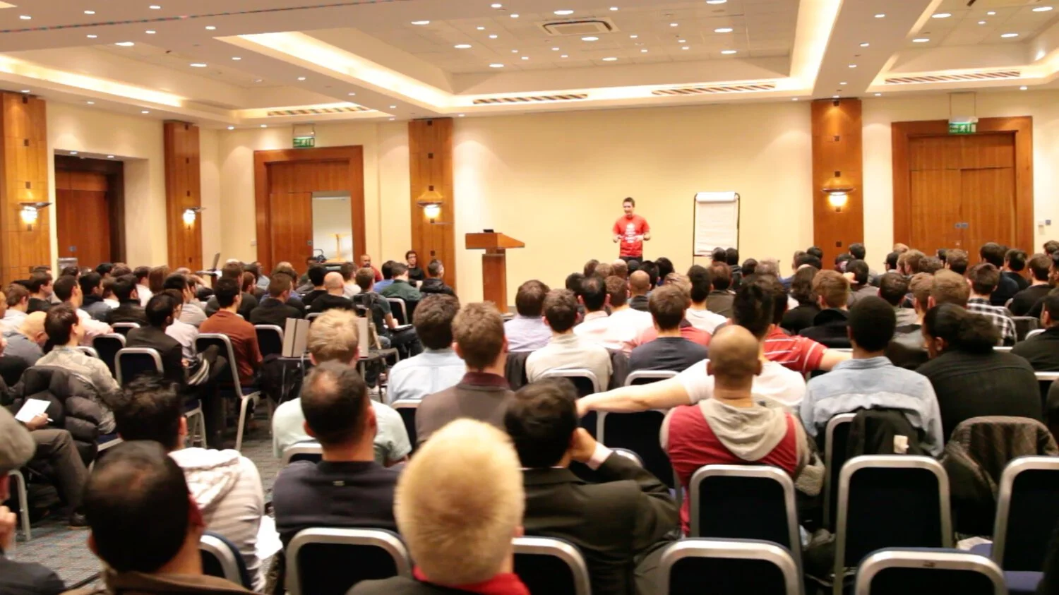 A large group of people attending a conference or seminar in a spacious conference room, with a presenter at the front near a podium and a flip chart.