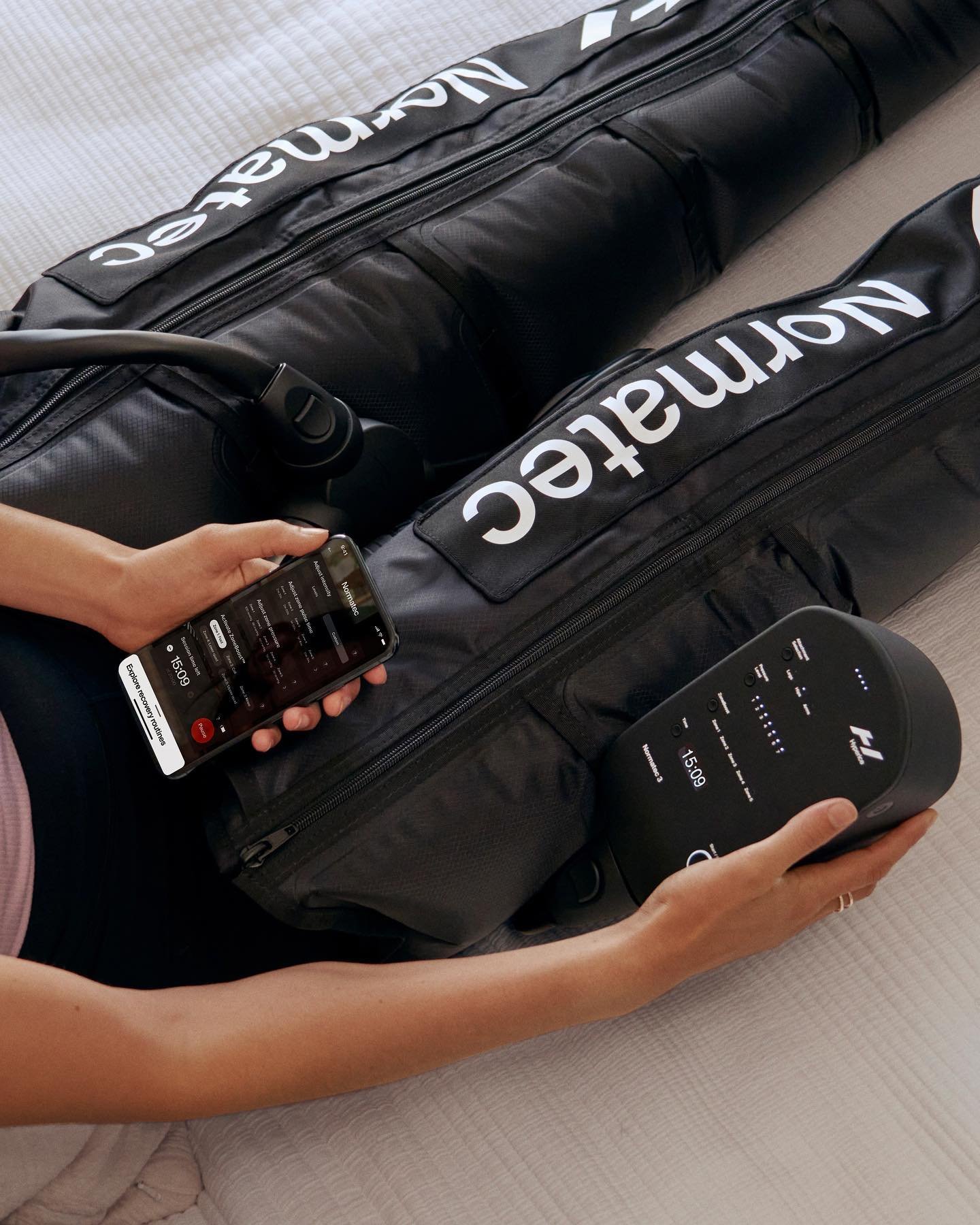 Person holding a mobile phone and a portable speaker while sitting next to two black bags with white 'NORMATEC' branding.