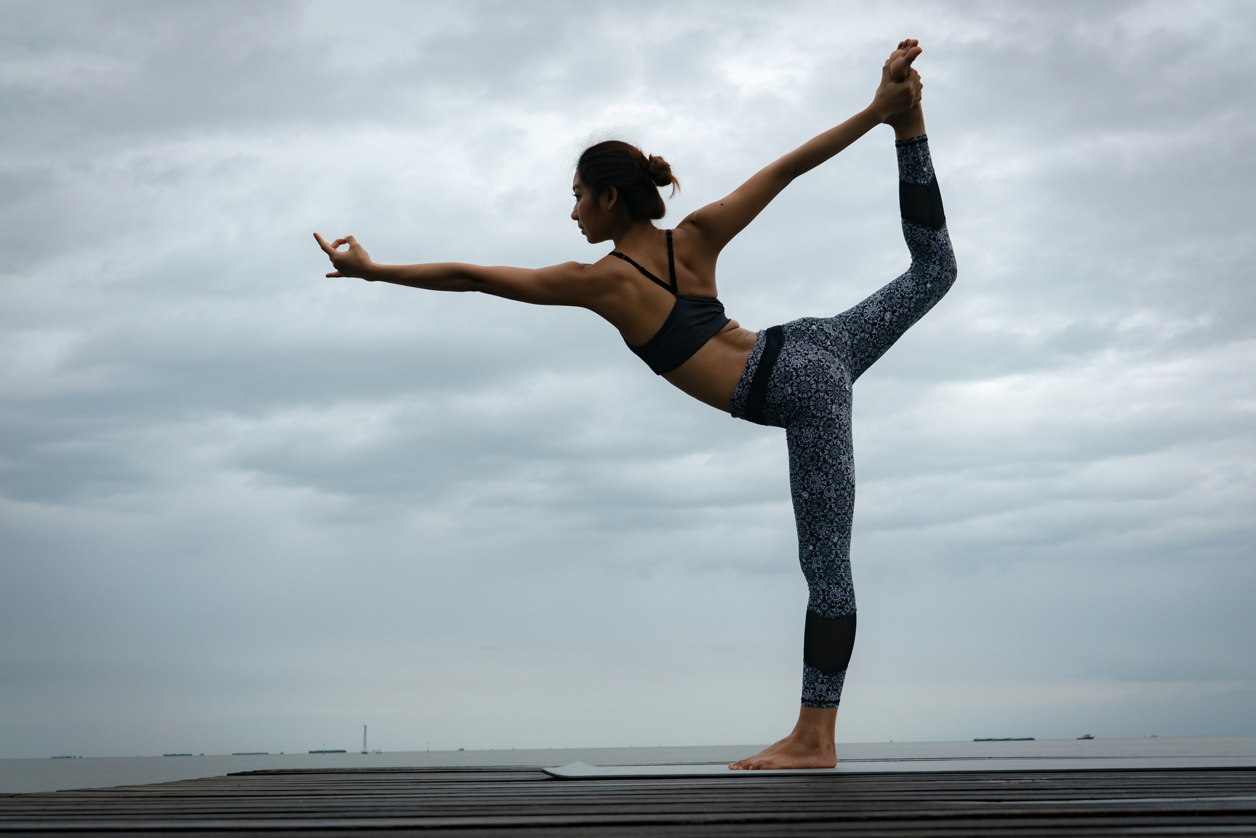 A woman practicing yoga outdoors on a wooden deck, performing a balancing pose with one leg lifted and arms extended, against a cloudy sky and distant ocean.