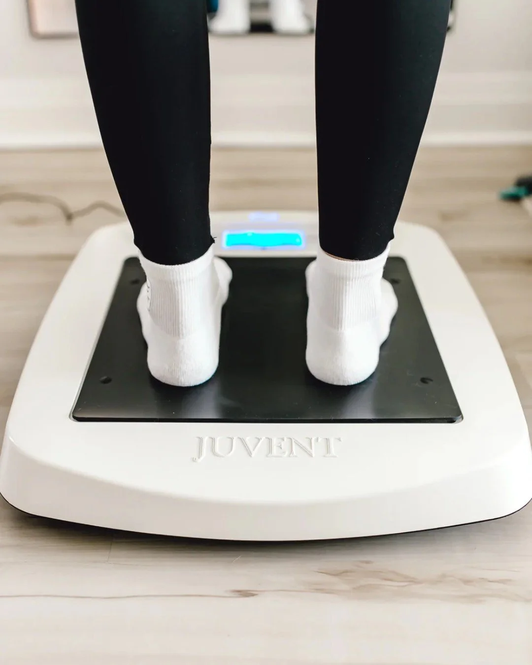 Person standing on a white digital scale, wearing black pants and white socks, in a room with a light-colored wooden floor.