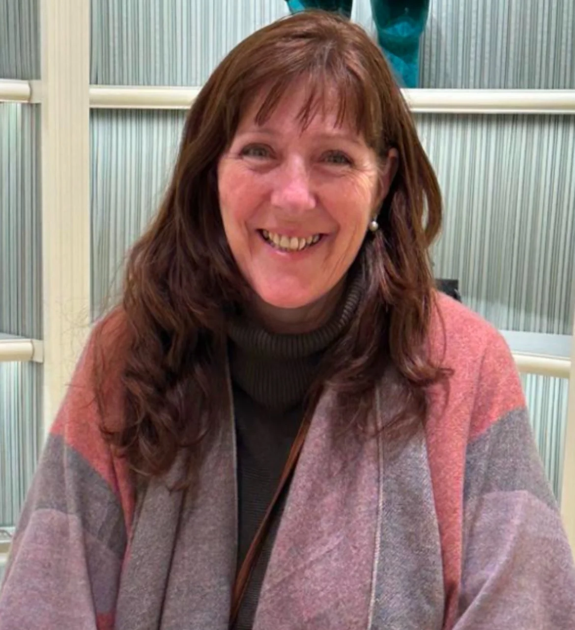 A smiling woman with reddish-brown hair, wearing a pink and gray patterned jacket and a dark turtleneck sweater, sitting in front of a background with vertical striped glass panels.