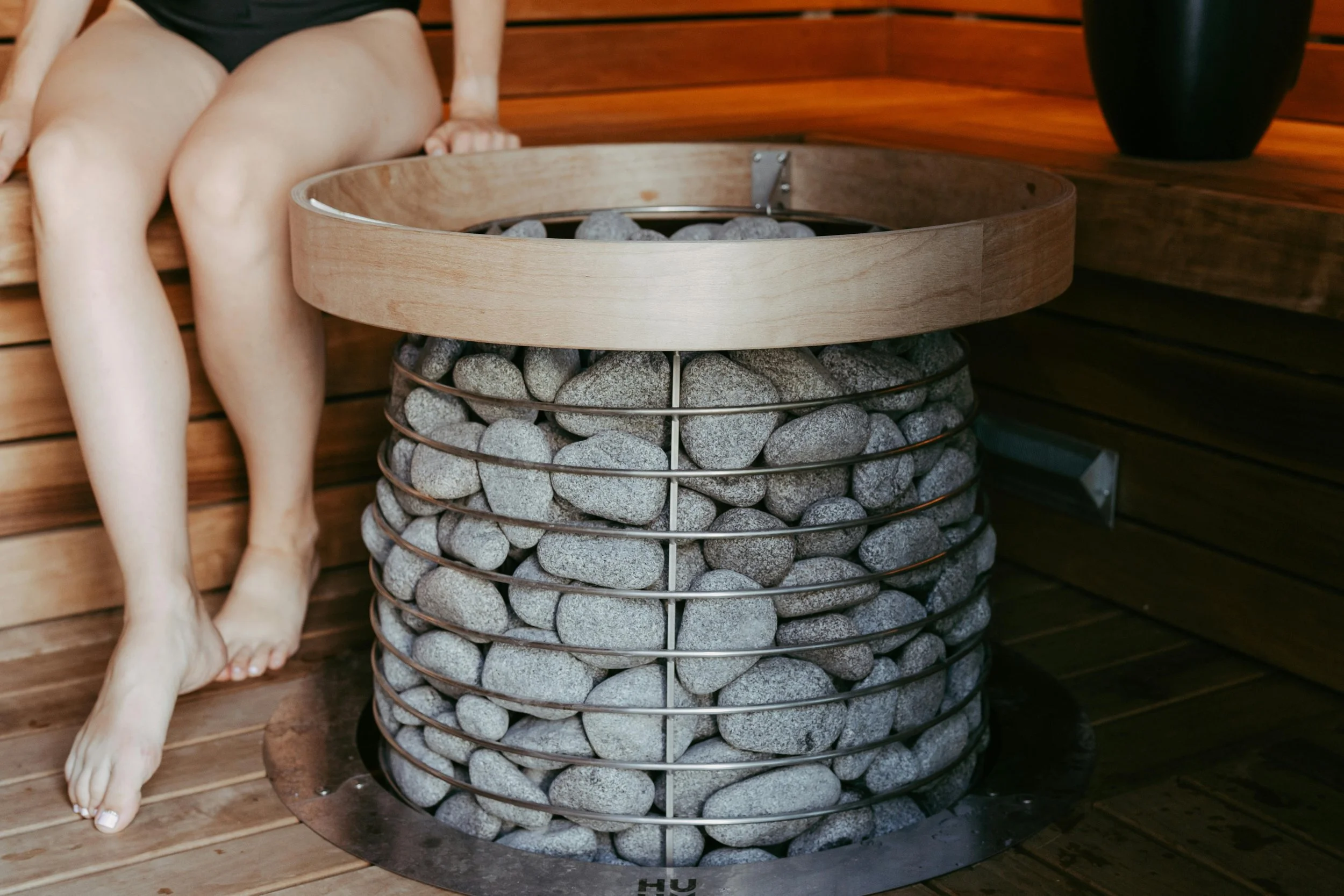 A wooden sauna with a round stone heater filled with stones. Part of a person sitting on a wooden bench is visible.
