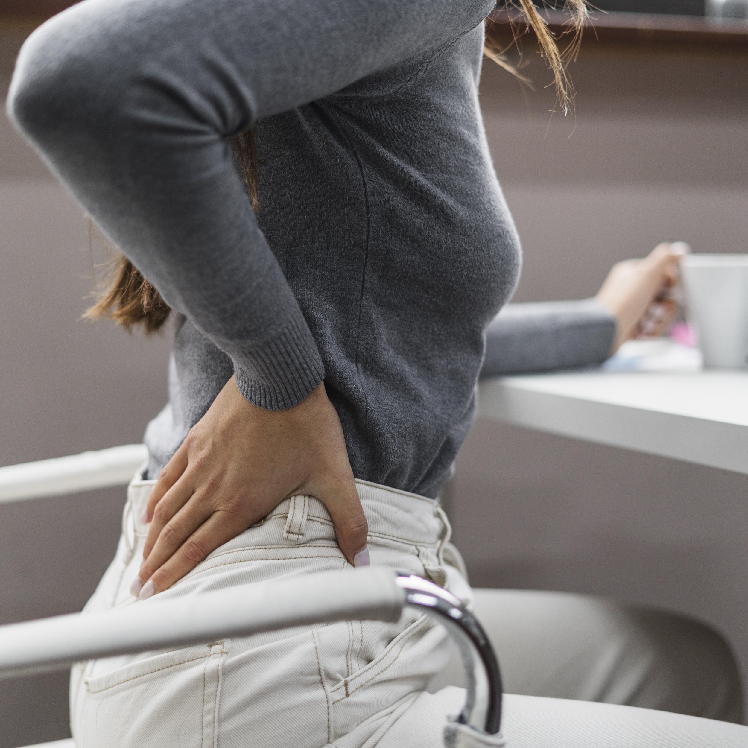 A person holding their lower back with one hand, demonstrating discomfort or pain, while sitting at a desk with a cup and other items.