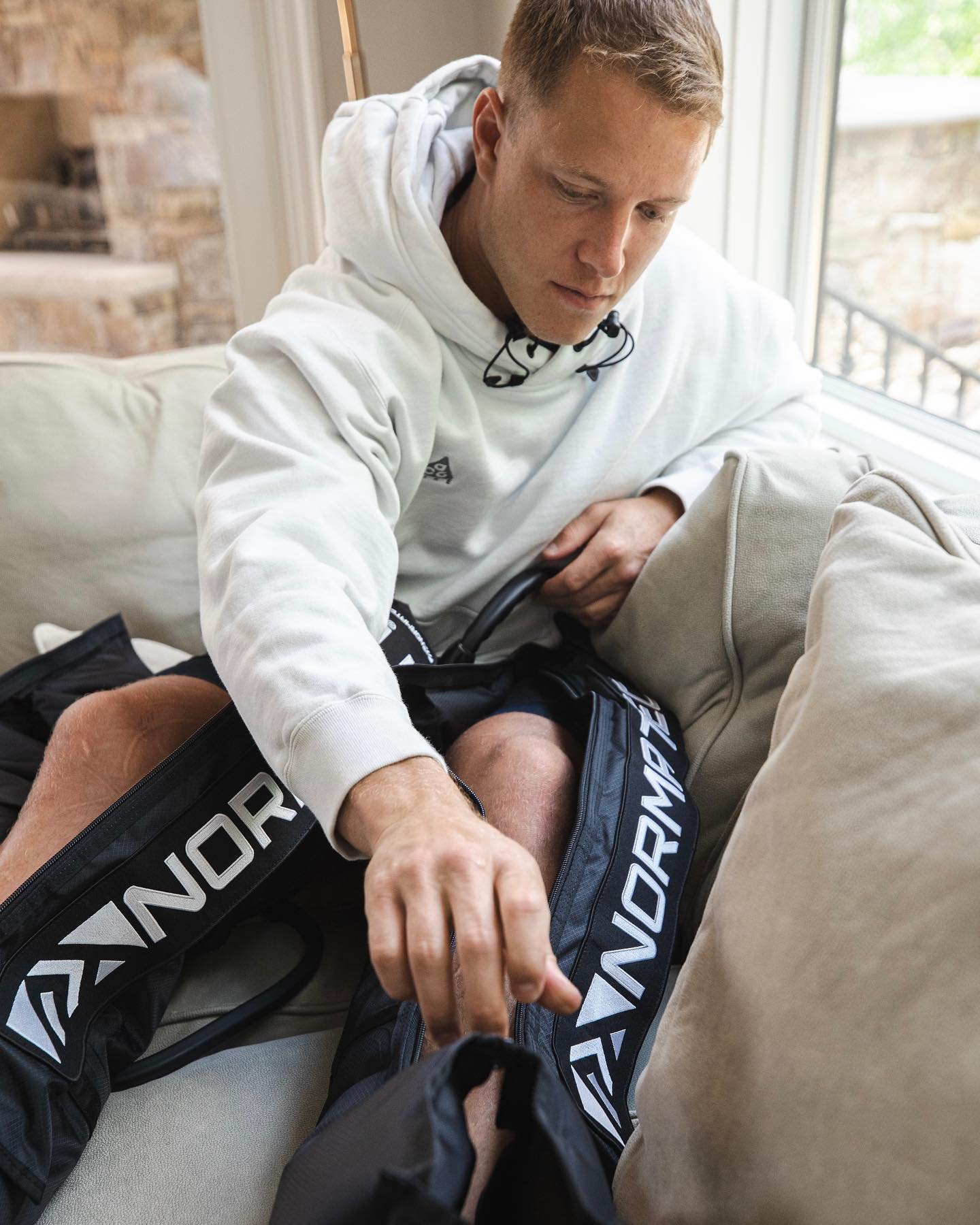 A man in a white hoodie sitting on a couch, reaching into a hockey bag with 'WARRIOR' branding, next to a beige cushion near a window.