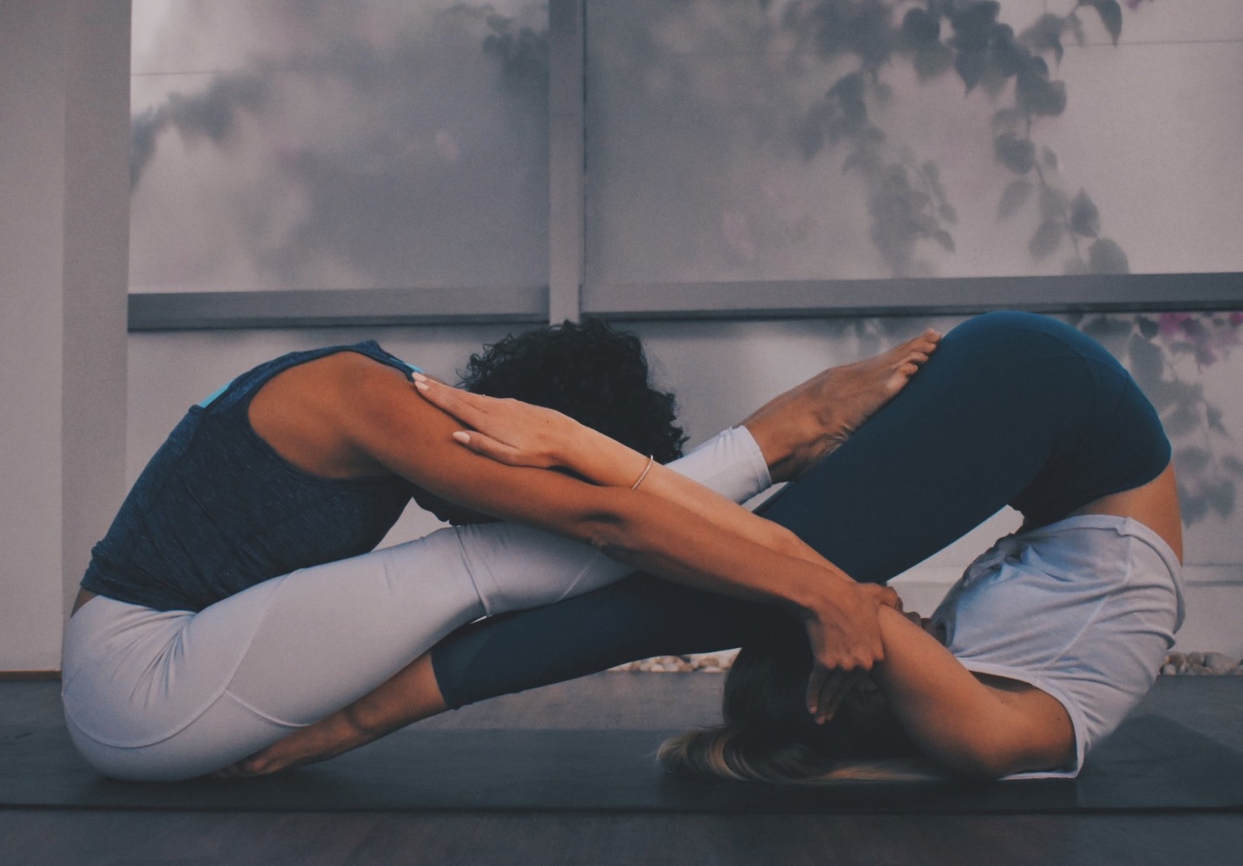 Two women practicing partner yoga on a black mat, one woman with curly hair and dark tank top supporting the other woman with long hair and a white t-shirt, both in a seated forward bend pose with arms extended, in a calm indoor setting with a backdrop of scenic clouds and tree silhouettes.