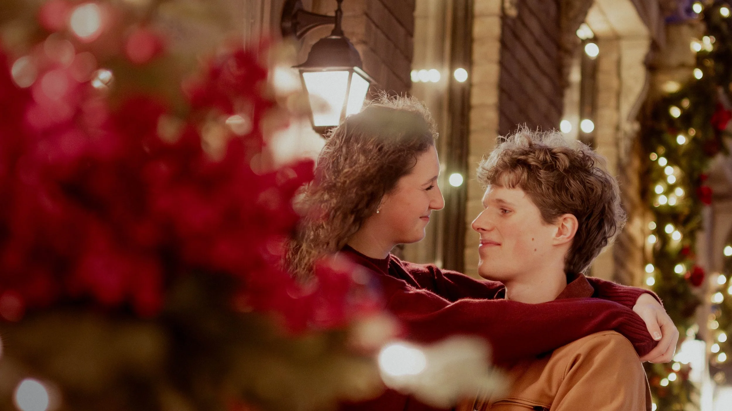 A woman and a young man sit close together, smiling at each other, in a cozy, festively decorated setting with Christmas lights and a decorated Christmas tree in the background, surrounded by blurred red holiday decorations in the foreground.