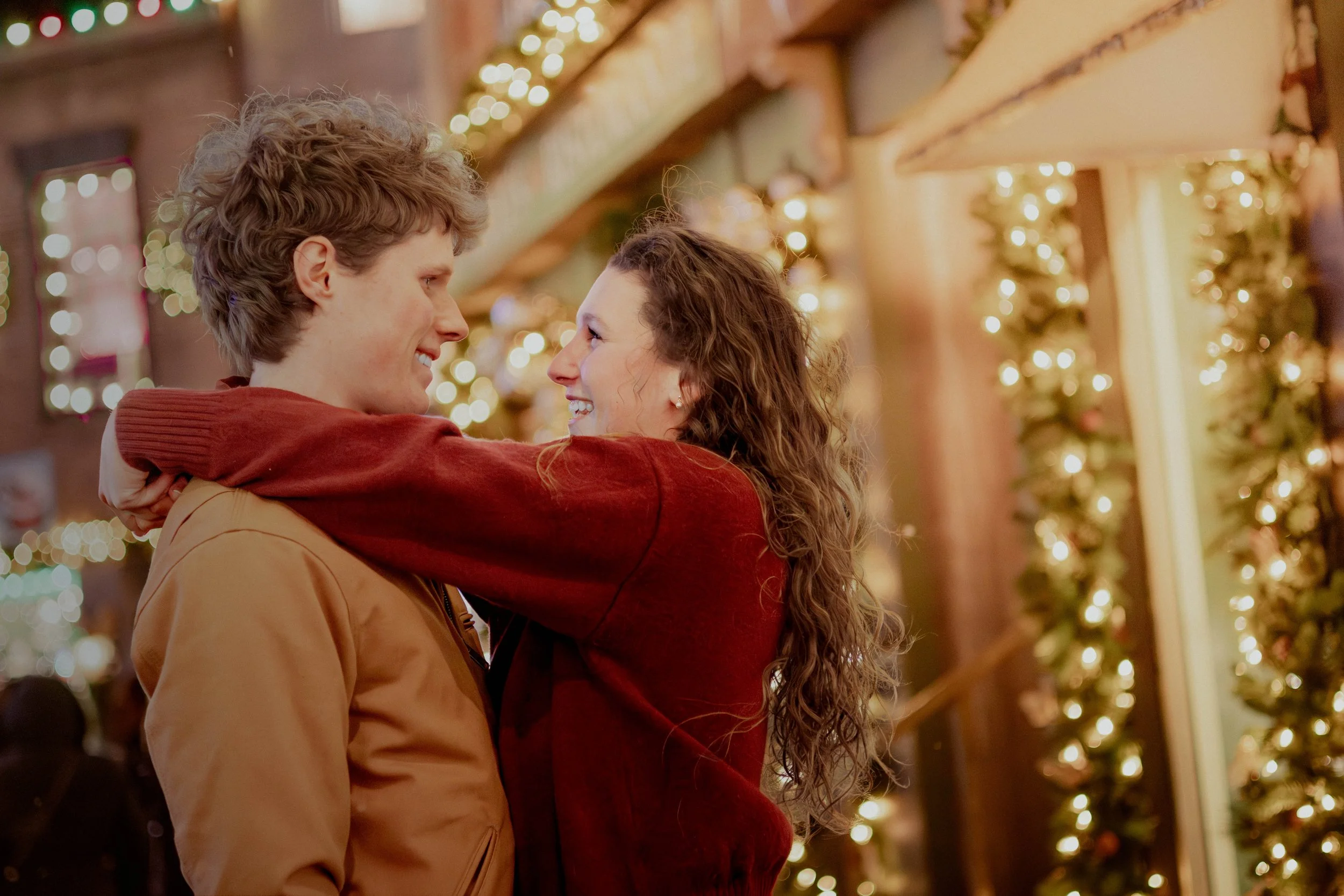 A couple hugging and smiling at each other in front of festive Christmas decorations with lights and greenery.