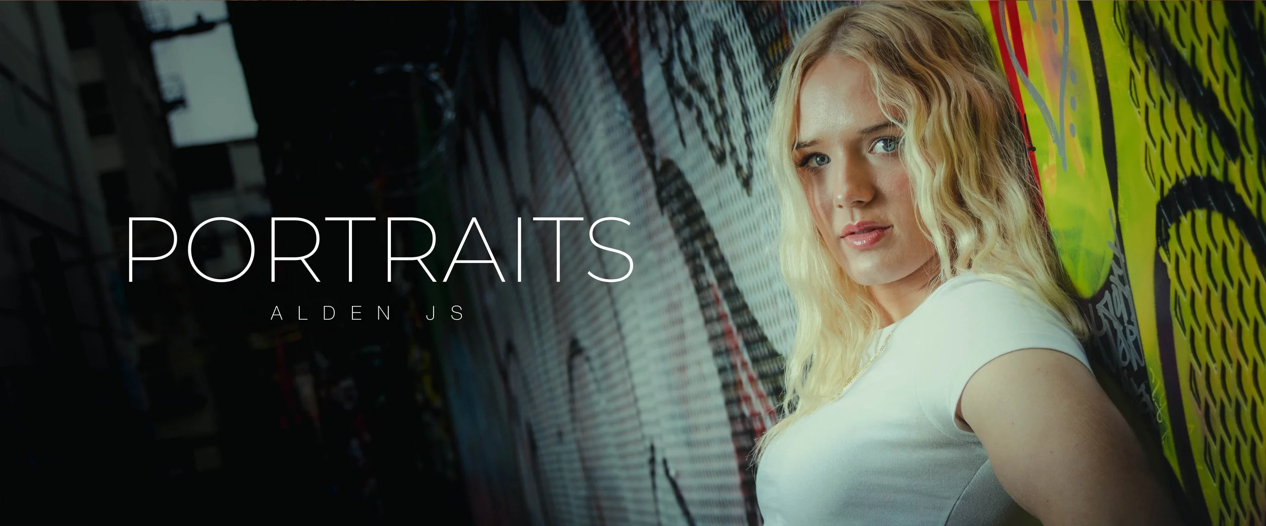 A young woman with blonde, curly hair wearing a white shirt, standing against a colorful graffiti wall.