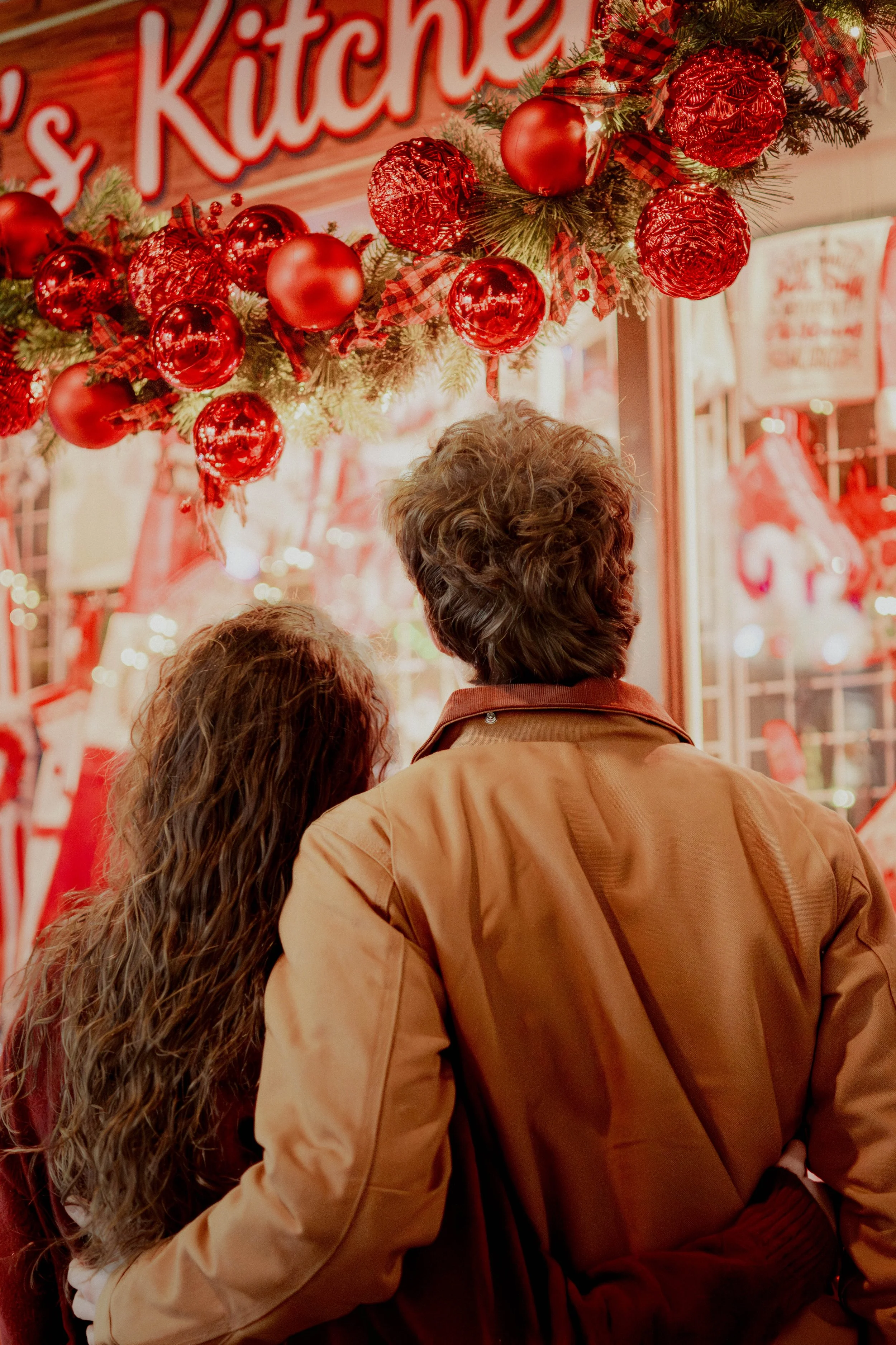 A couple with their backs facing the camera, standing in front of a Christmas-themed display with red ornaments and decorations at night.