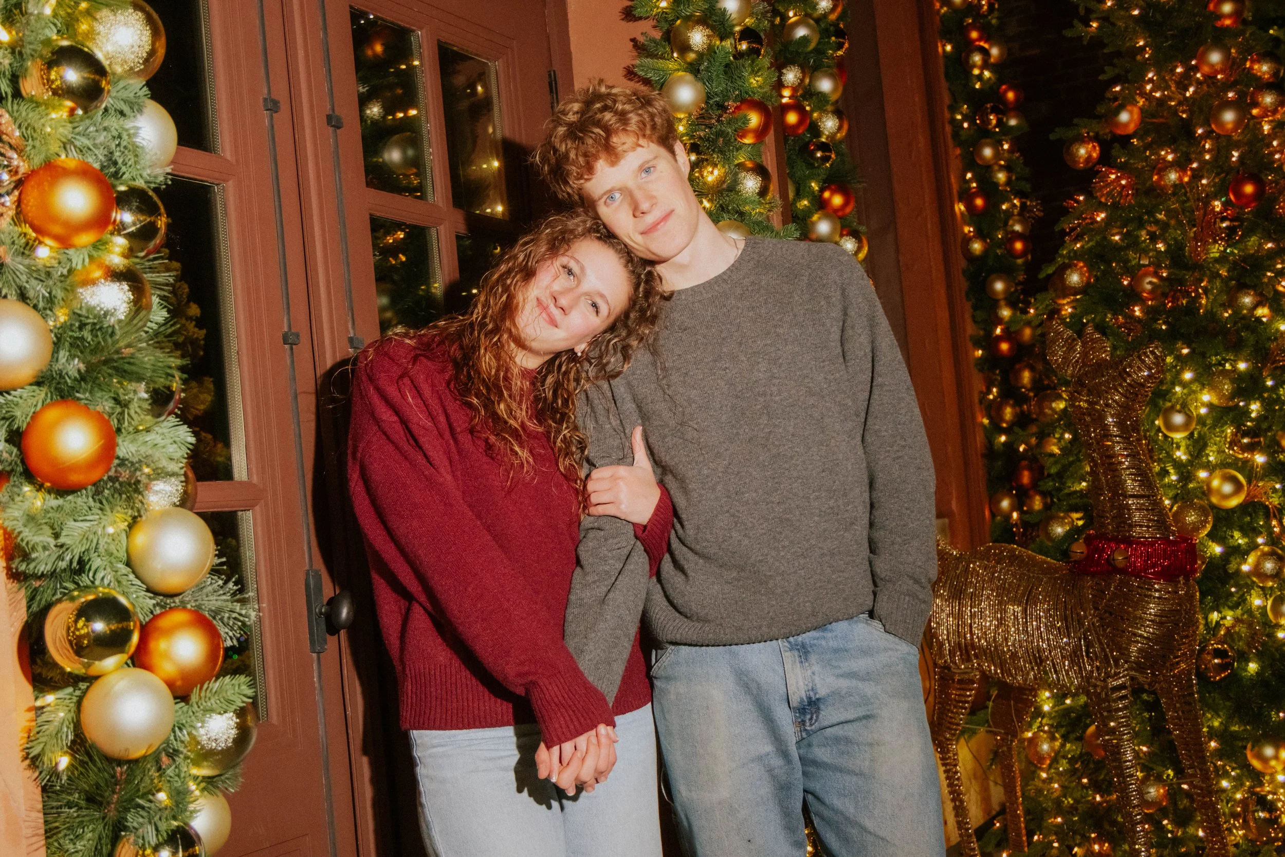 A young couple standing close together, holding hands and smiling, in front of a decorated Christmas tree with gold and red ornaments, illuminated lights, and a reindeer figurine, during the holiday season.